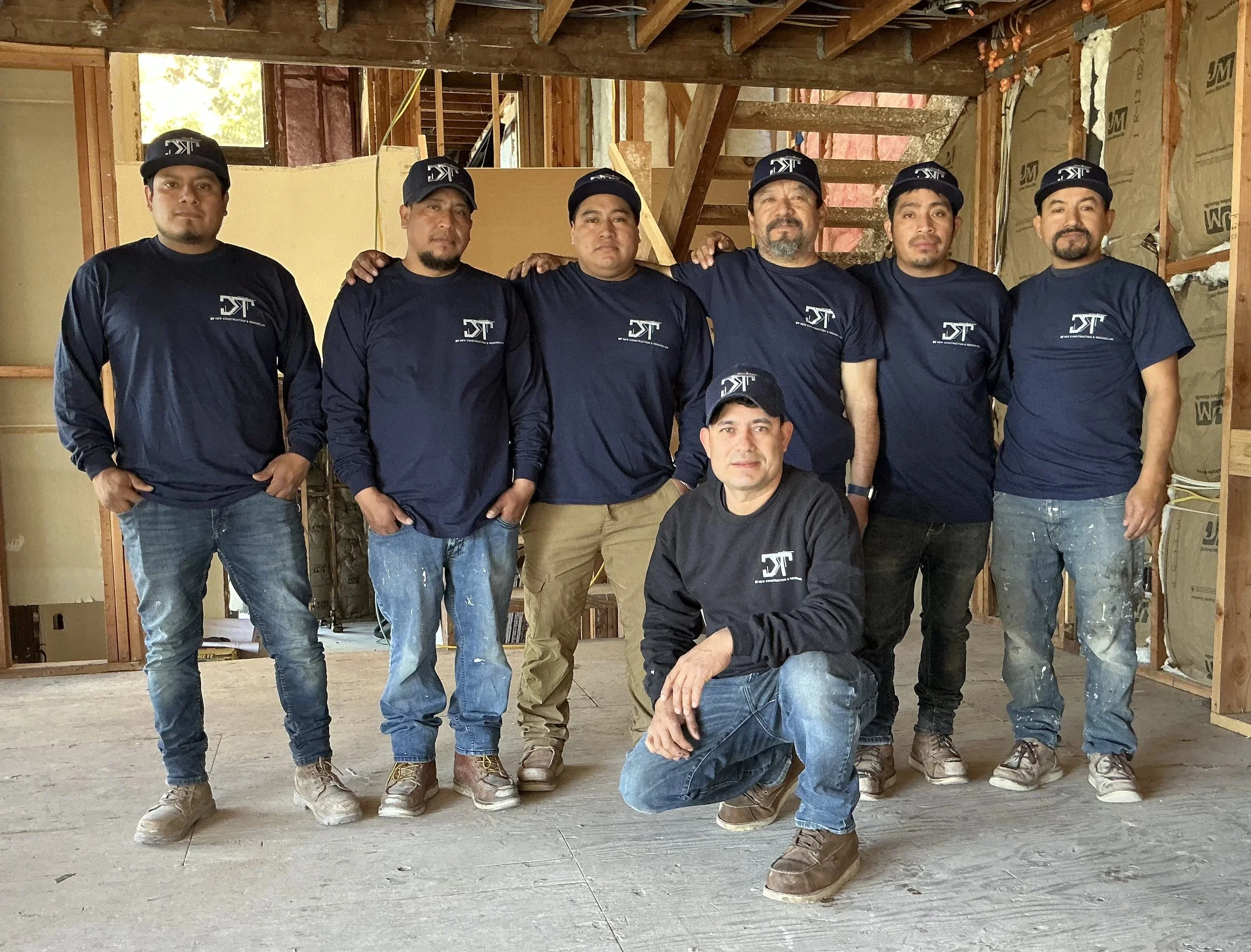A group of seven construction workers inside a building under construction, posing for a photo. They are wearing matching navy blue shirts and caps with a logo, and are standing or kneeling inside the unfinished structure with exposed wooden framing.