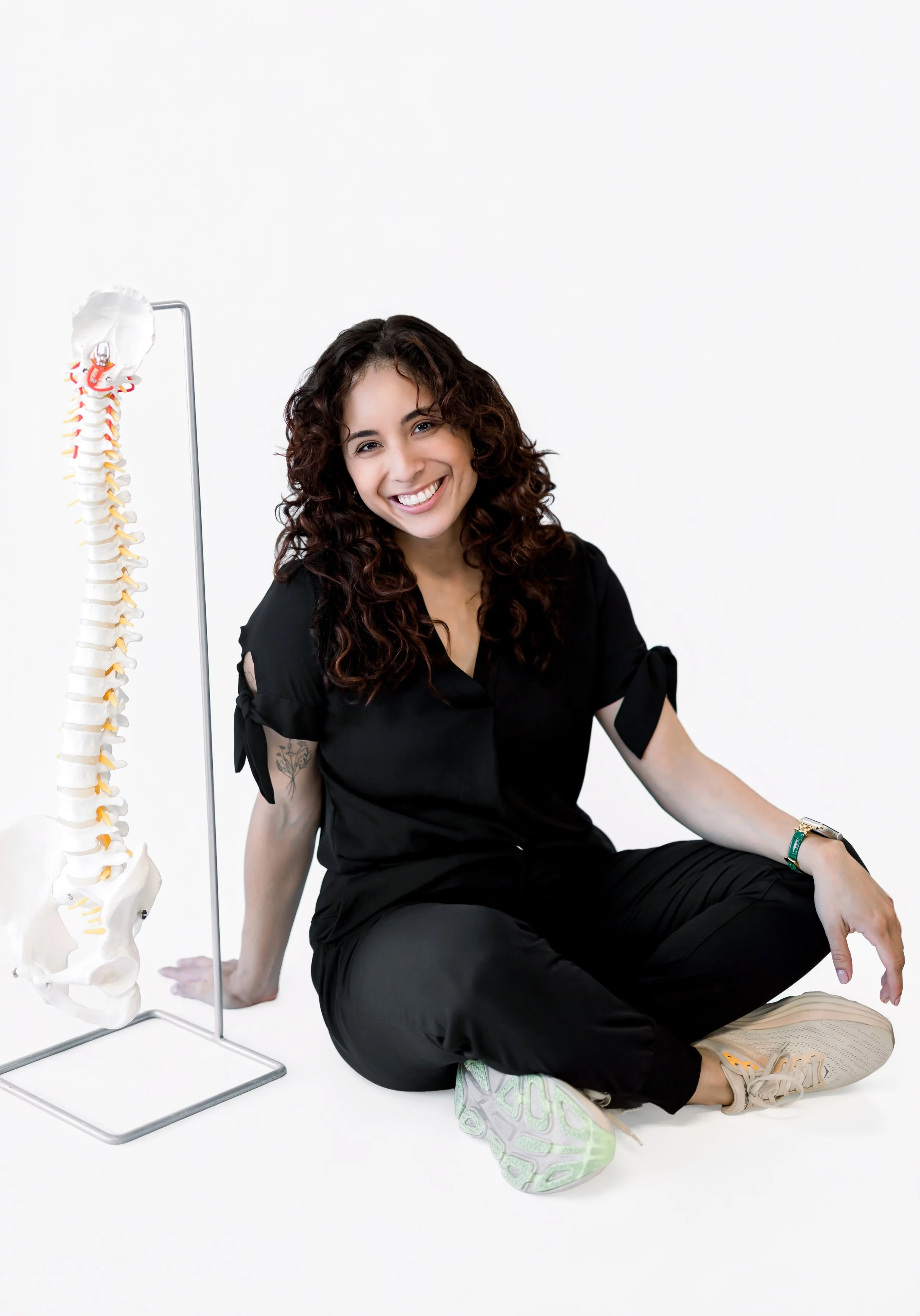 A woman with curly brown hair, wearing a black shirt and black pants, sitting on the floor with a model of a human spine on a stand next to her, smiling at the camera, against a plain white background.