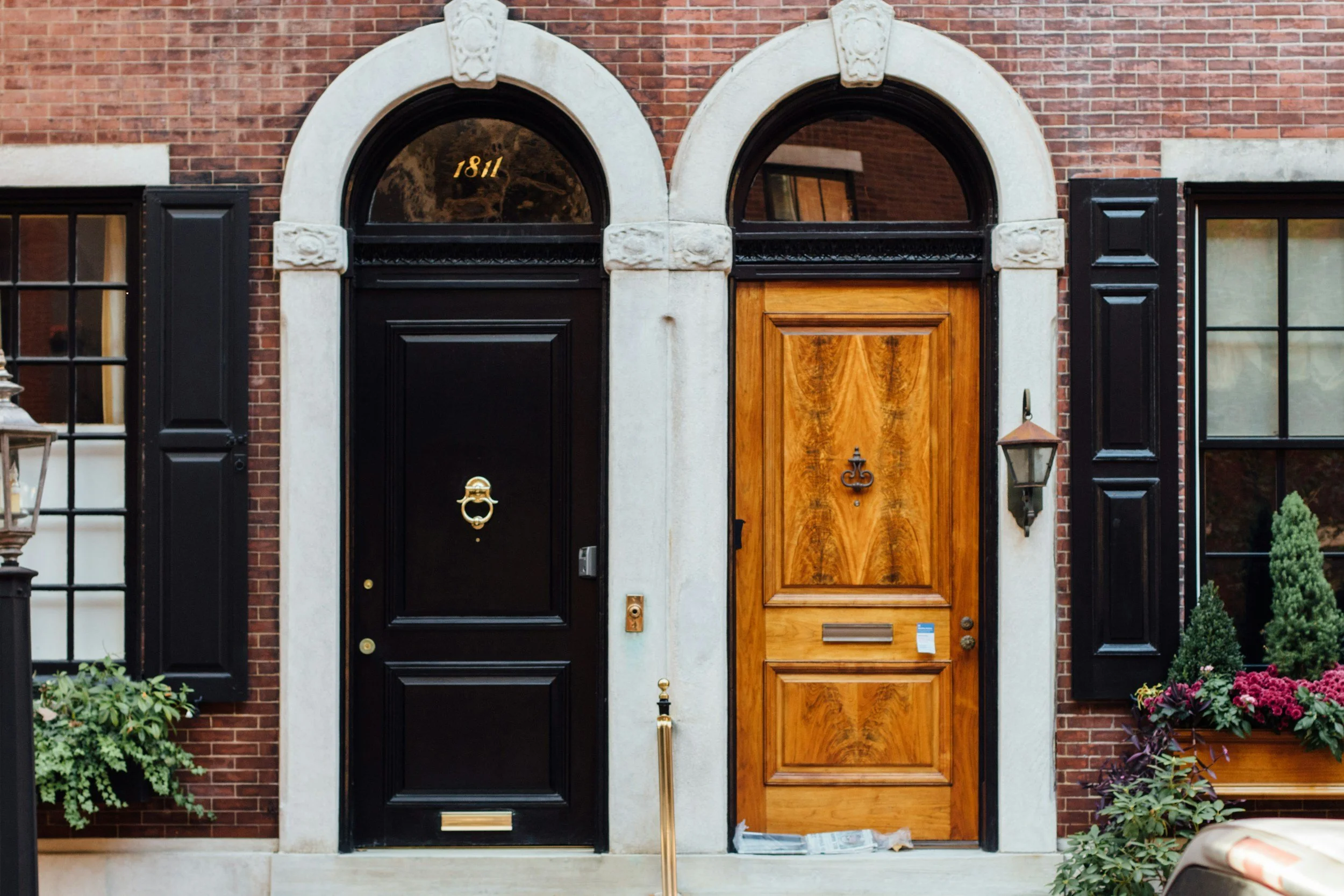 Close-up of two adjacent front doors, one black and one wood, in a brick building. The black door has a brass knocker; the wood door has a decorative handle and mail slot. Above the doors are arched transom windows, with house number 1811 in the black window.