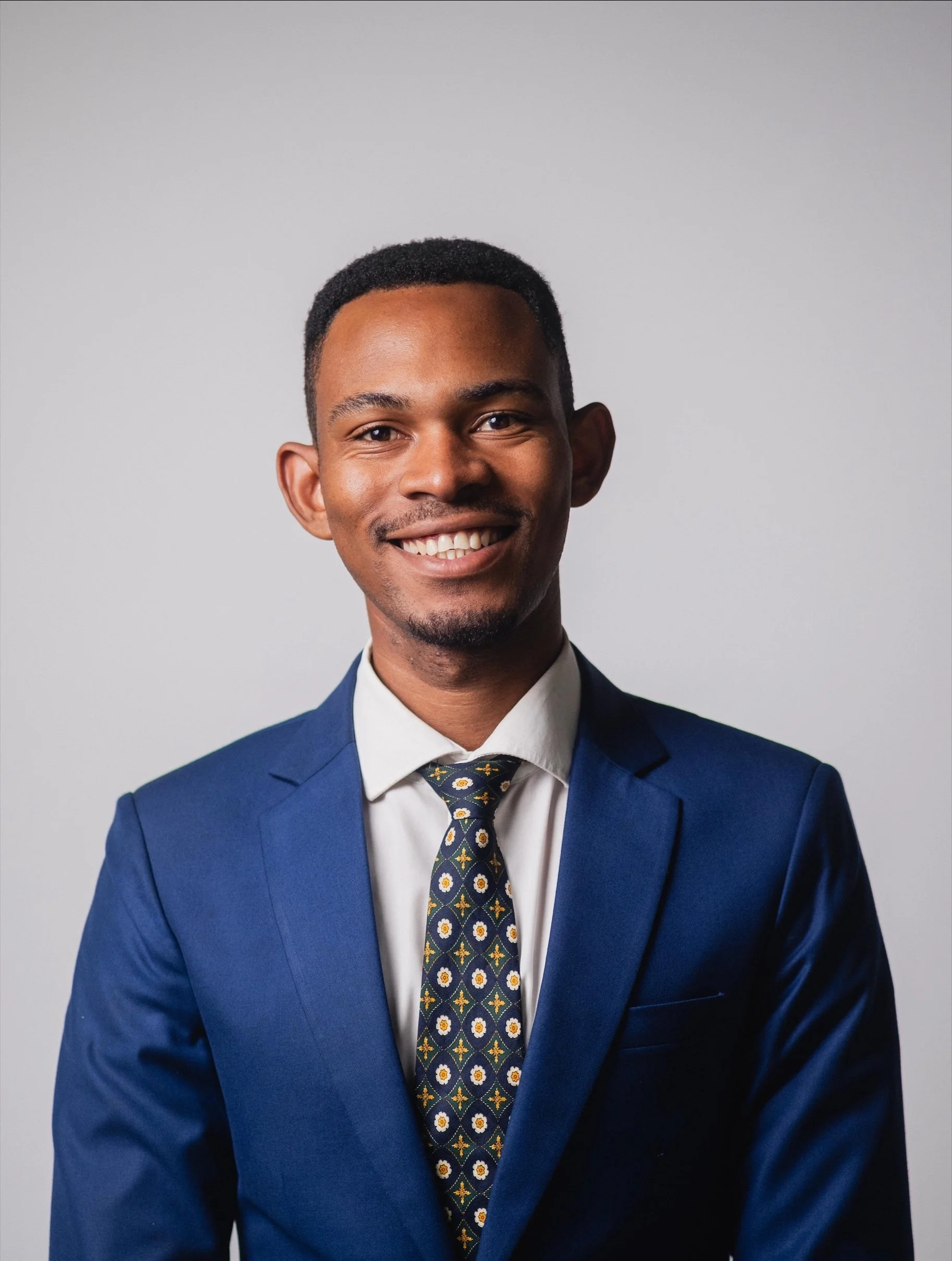 A young man in a navy suit, white shirt, and patterned tie smiling against a plain light gray background.