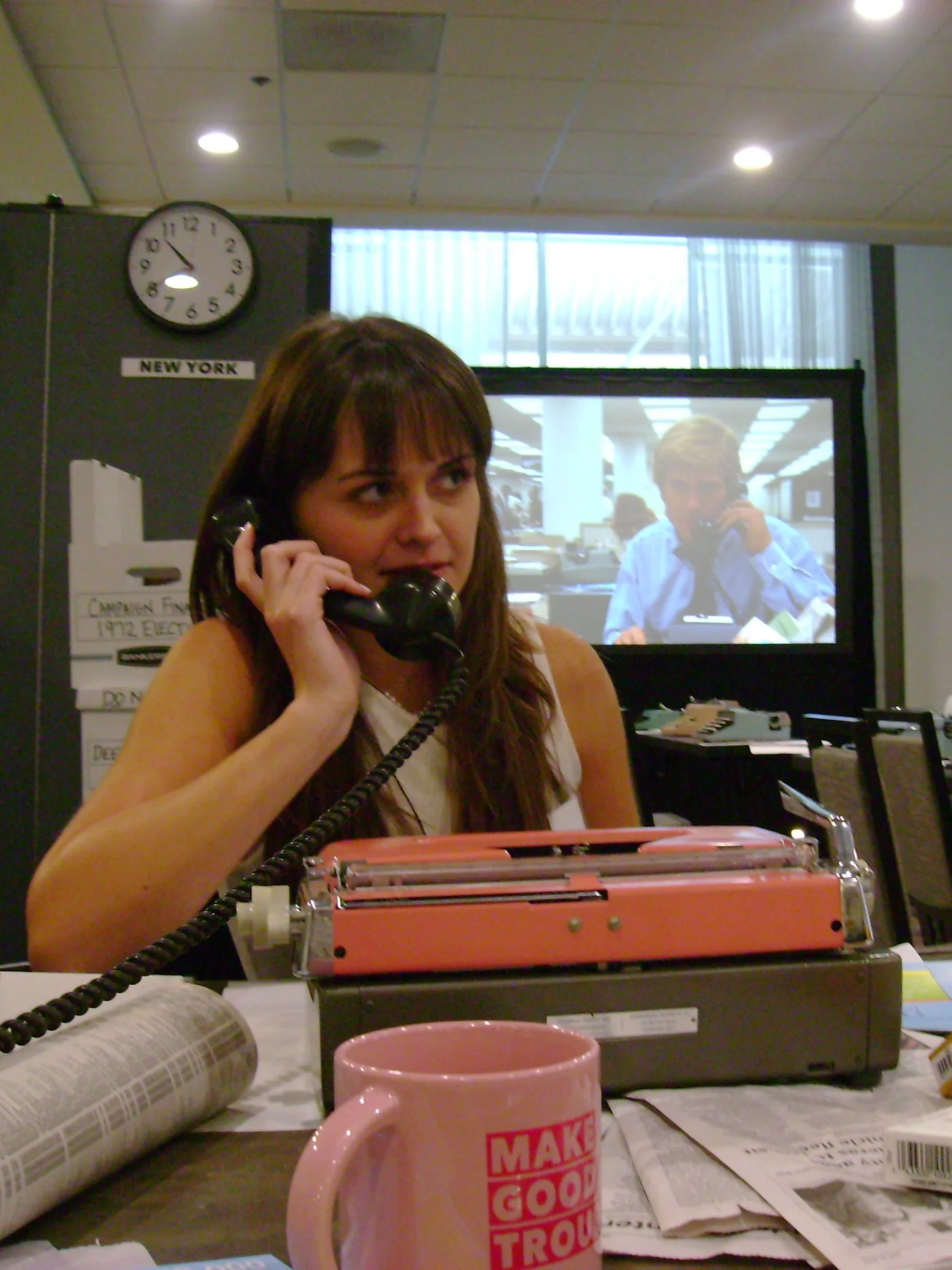 A woman with long brown hair talking on a black landline phone at a desk. In front of her, there's a pink mug and some papers. In the background, a television screen shows a man talking on the phone in an office. A clock on the wall shows approximately 9:09. The setting appears to be an office with a cubicle and overhead lighting.