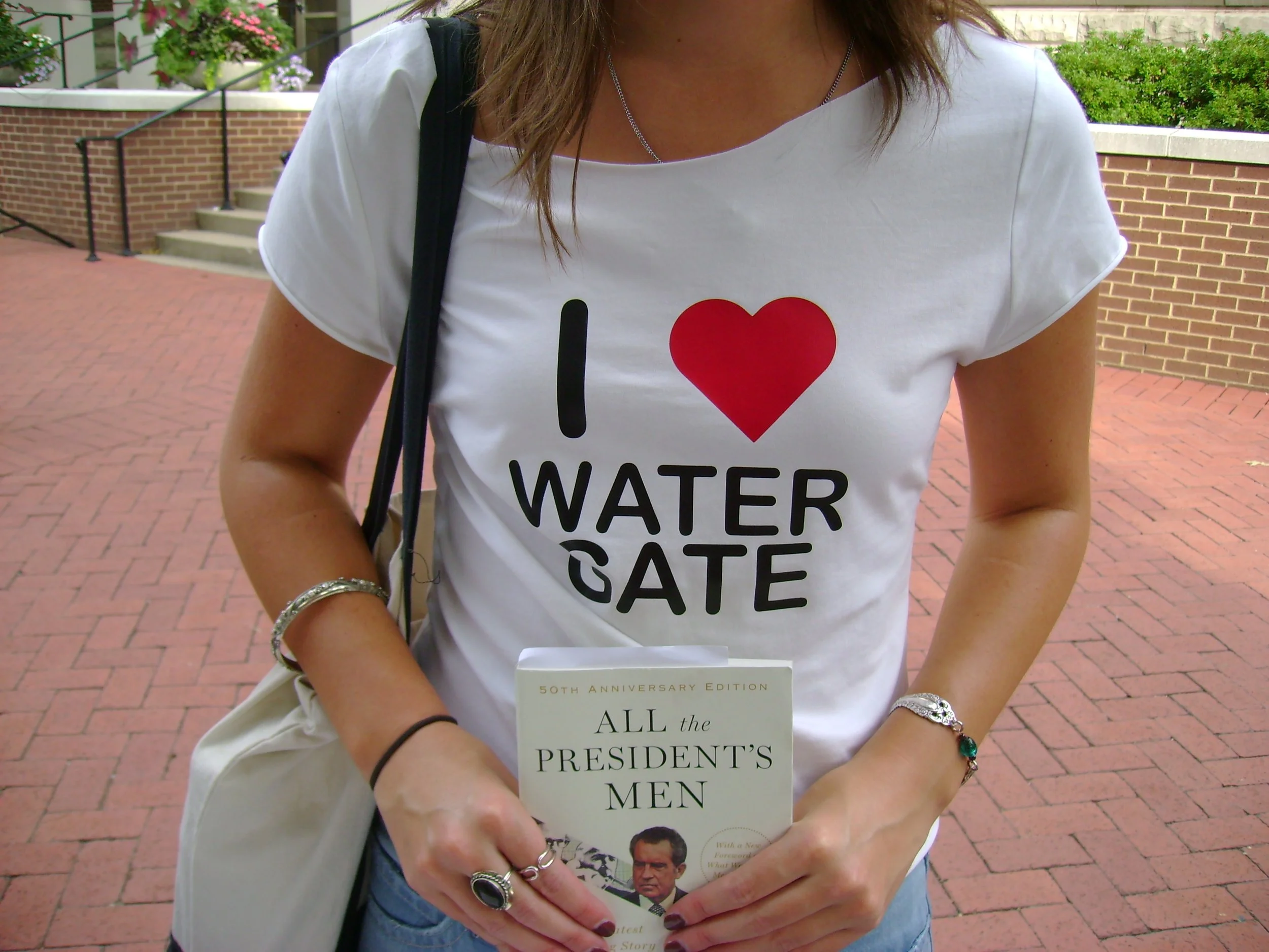 Woman wearing a white T-shirt with the text 'I ❤️ Watergate' printed on it, holding a book titled 'All the President's Men' outside on a brick walkway.