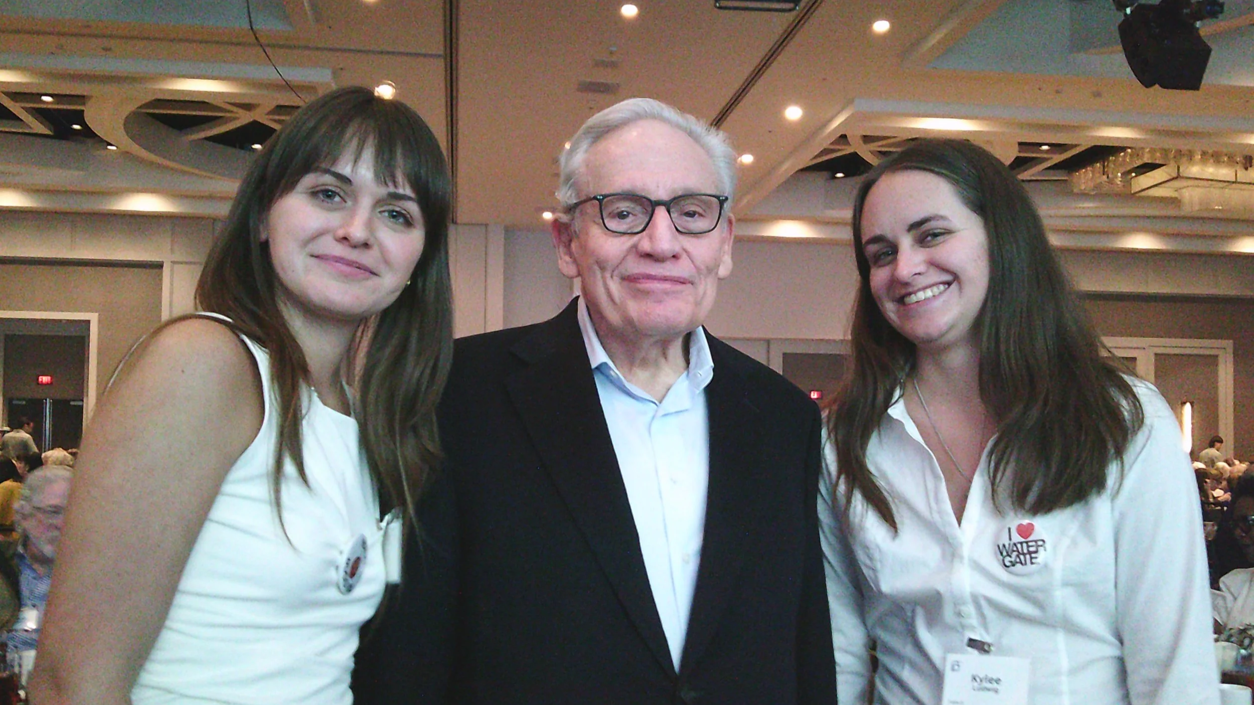 Three people standing together indoors at an event, with the man in the middle and two women on either side, all smiling.