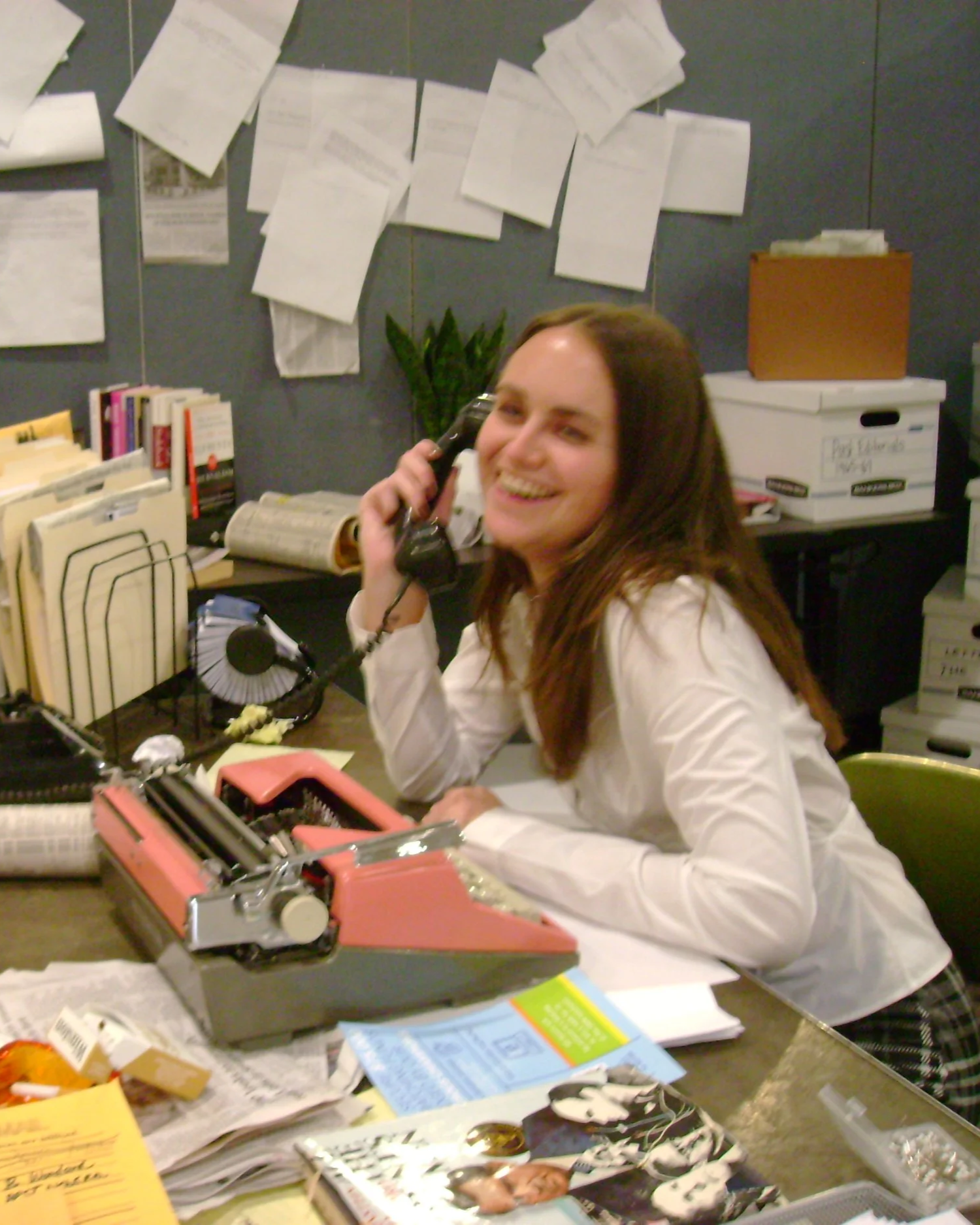 A woman with long brown hair in a white shirt smiling while talking on a black telephone in an office with papers, books, and office supplies on the desk.