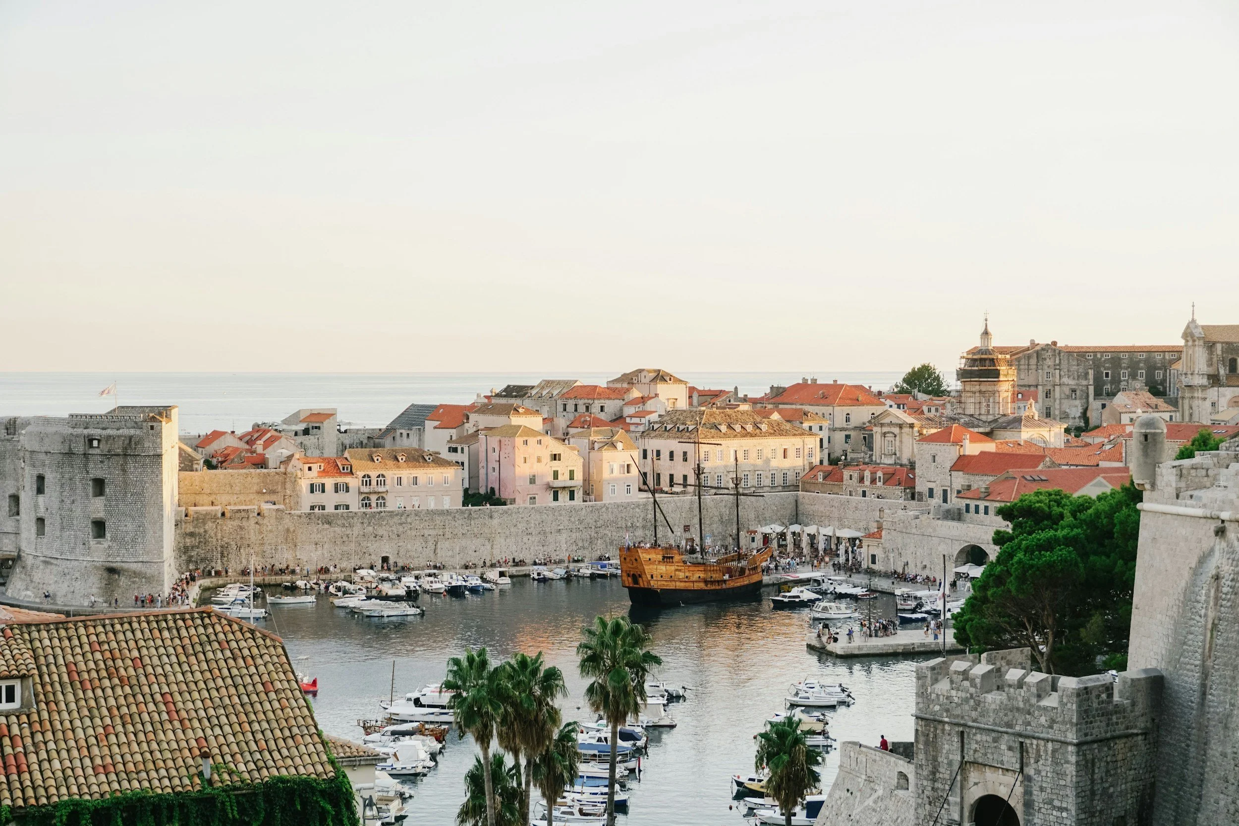 A scenic view of a historic coastal city with stone walls, a harbor full of boats including an old wooden pirate ship, colorful buildings with red-tiled roofs, and lush green palm trees.