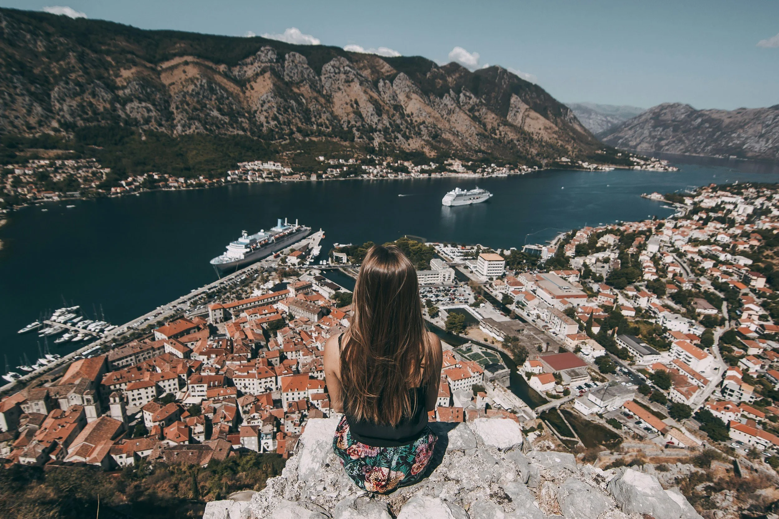 A woman sitting on a rocky ledge overlooking a coastal town with a harbor, boats, and a large cruise ship, with mountains in the background.