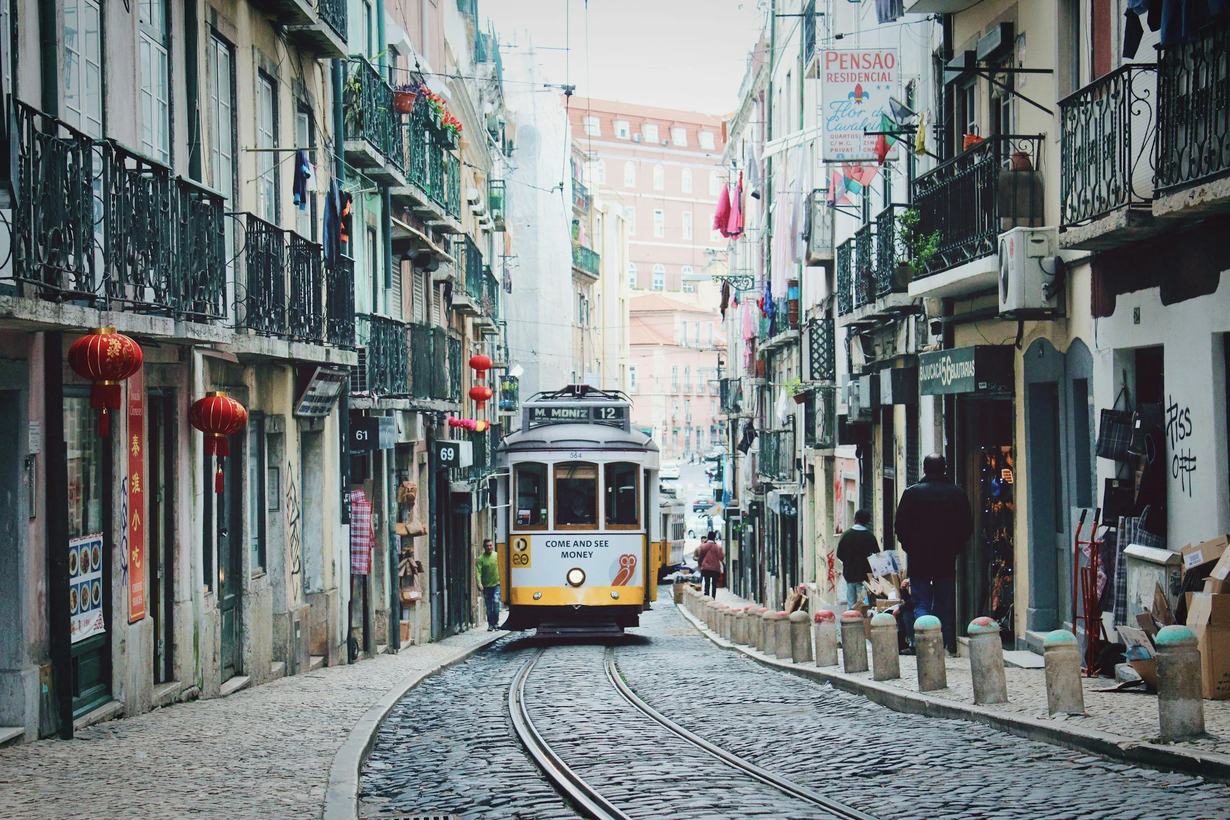 A street scene with a tram approaching, cobblestone road, and buildings with balconies on either side. Decorative lanterns hang along the street, and pedestrians are walking by.
