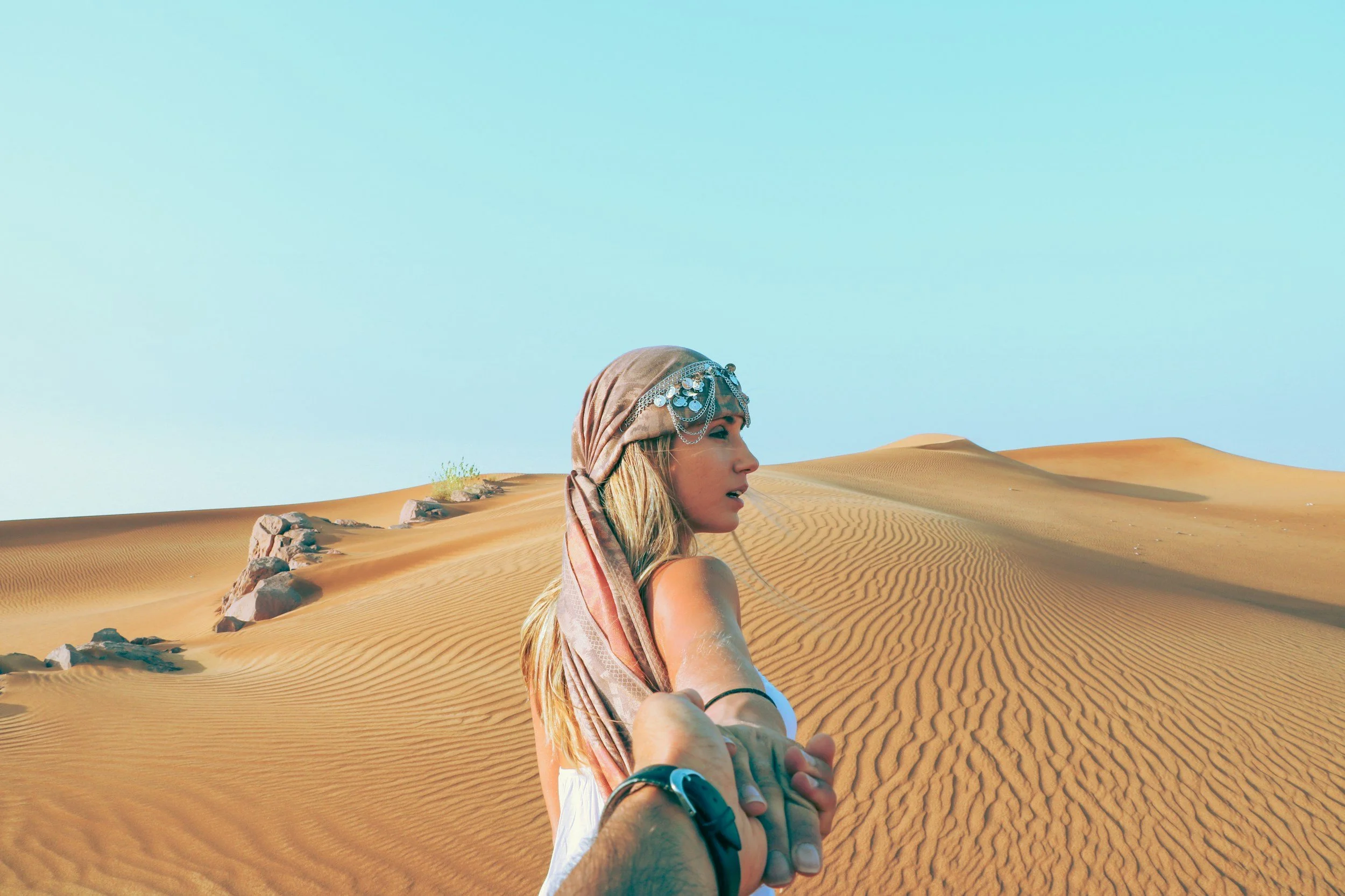 A woman wearing a pink bandana in a desert holding a man's hand looking backwards at him