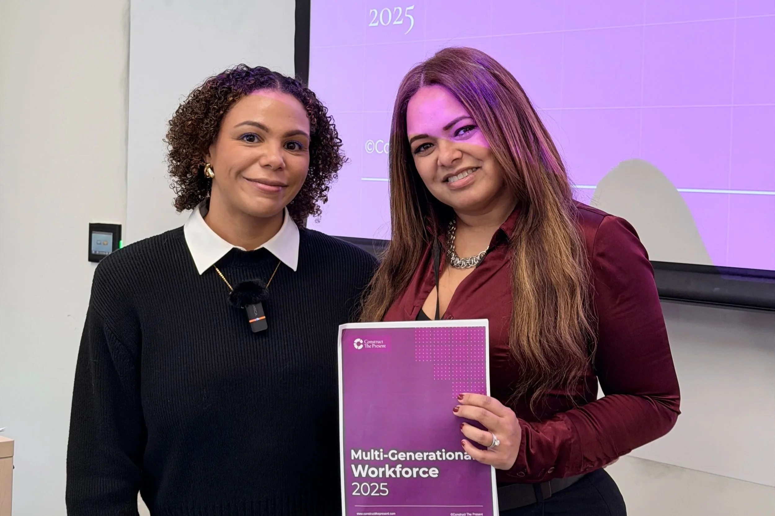 Two women standing side by side, smiling at the camera. One woman on the right holds a purple booklet titled 'Multi-Generational Workforce 2025', and is wearing a burgundy shirt with jewelry. The other woman on the left is dressed in a black sweater with a white shirt underneath and a small black microphone clipped to her outfit. Behind them is a large screen displaying a purple background with some text.