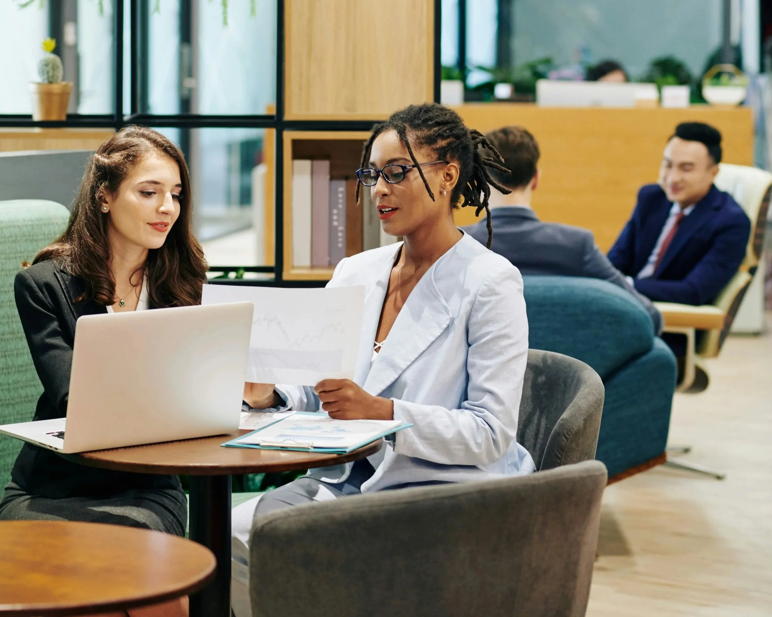 Two women work together at a small round table in an office lounge. One woman, with long brown hair, is looking at a laptop, while the other woman, with glasses and dreadlocks, is holding a document and speaking. In the background, three men are seated on couches engaged in conversation.