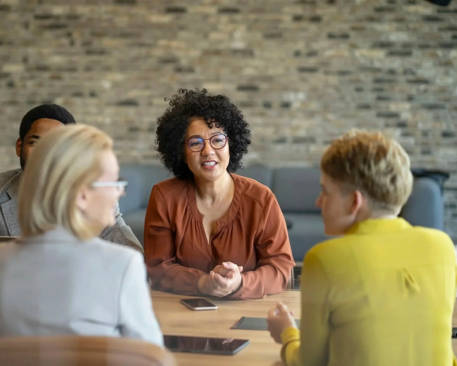 Group of diverse professionals in a meeting room, engaged in a discussion, with a woman speaking and others listening, in a modern office with a brick wall background.