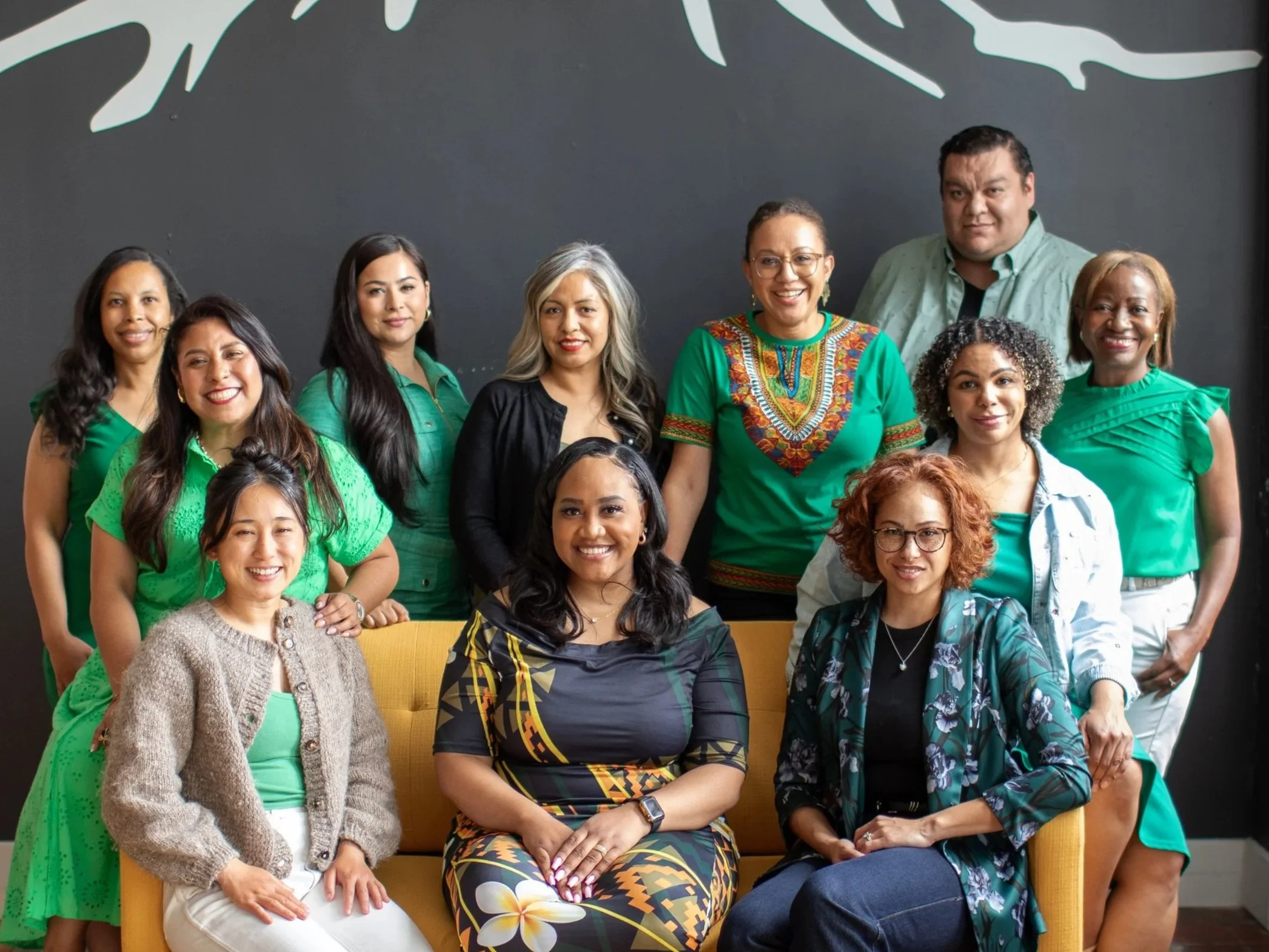A diverse group of twelve women and one man posing for a photo in front of a black wall with white abstract patterns. They are dressed in casual and colorful clothing, some smiling and some with neutral expressions.
