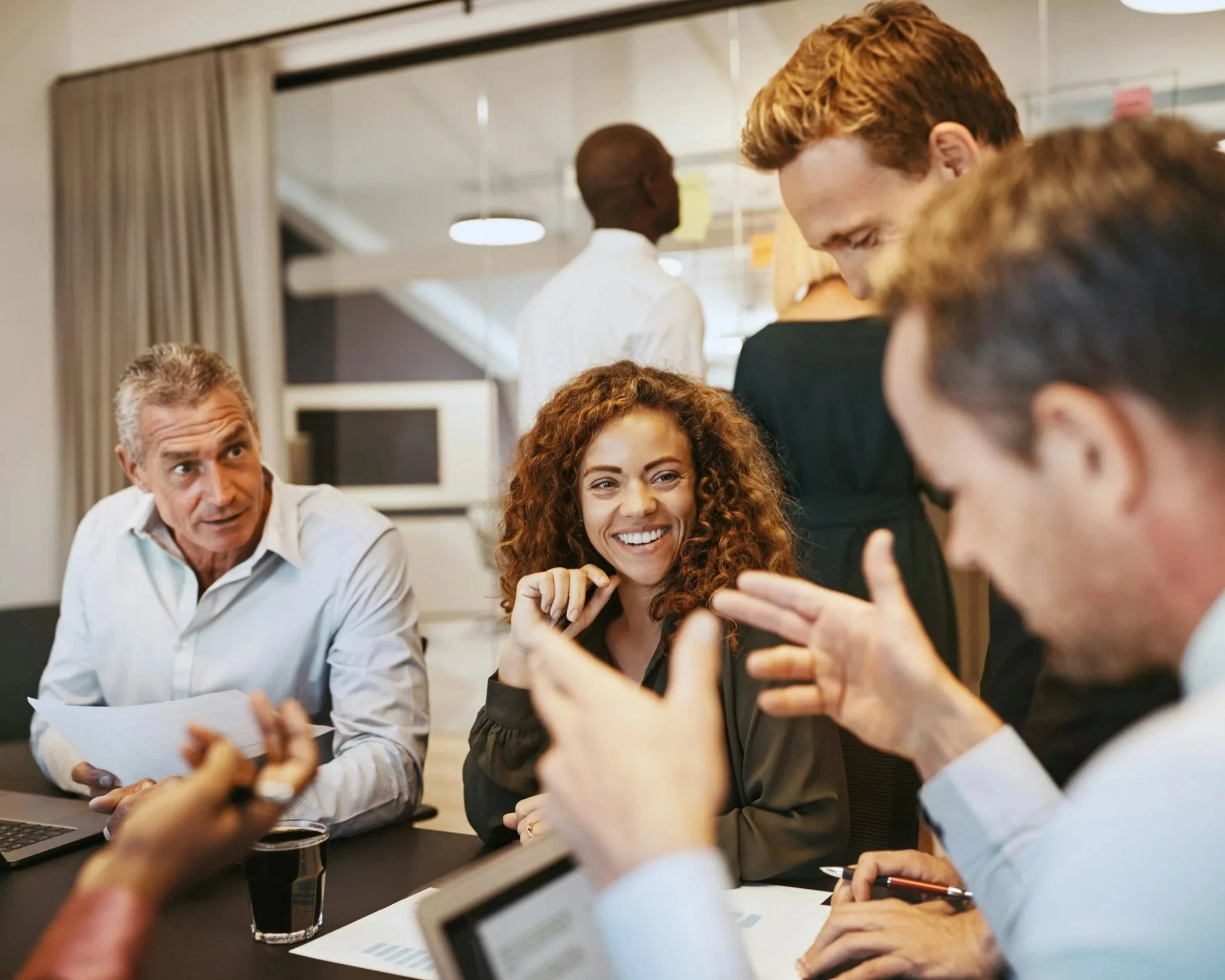 People in a business meeting or discussion, with men and women talking and smiling around a conference table in an office setting.