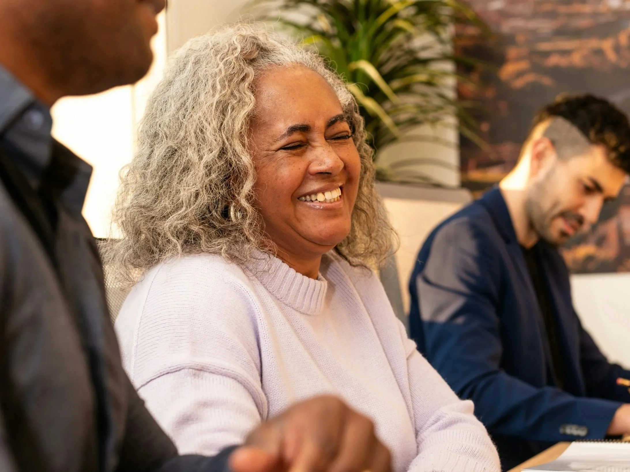 A diverse group of people sitting at a conference table, with a woman smiling and laughing, wearing a light pink sweater, and two men nearby, one on each side.