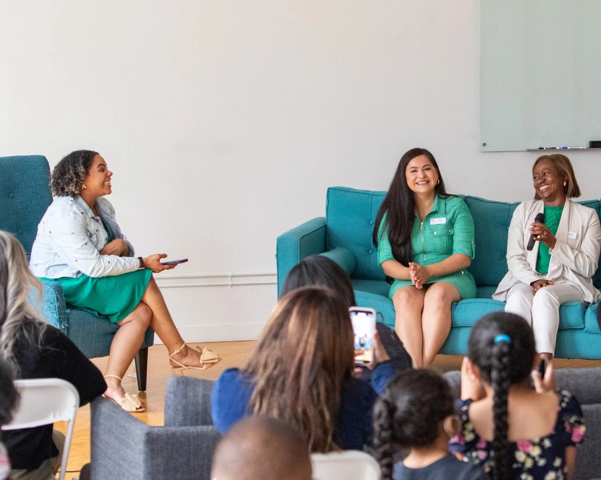 Three women sitting together on a teal couch and chair, smiling during a panel discussion or event, with audience members in the foreground.
