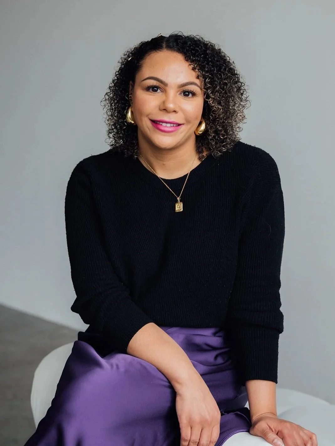 A woman with curly dark hair wearing a black sweater, purple skirt, gold earrings, and a gold necklace with a letter A pendant, sitting on a white chair against a gray background.