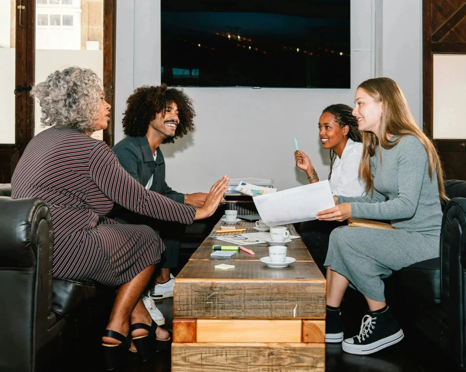 Five diverse women having a friendly meeting at a wooden table in a cozy cafe, engaged in conversation and smiling.
