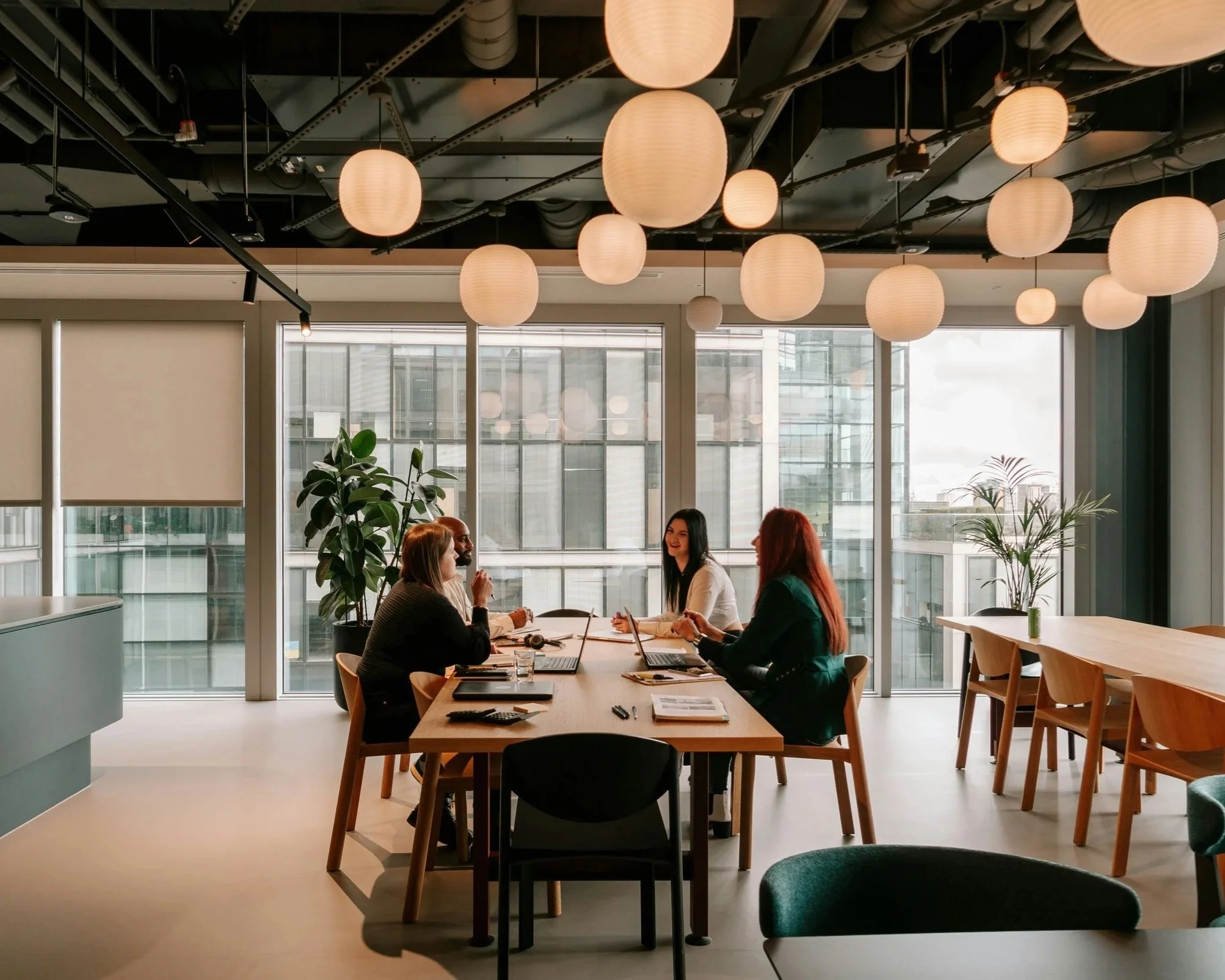 Four people in business attire sitting at a meeting table in a modern office with large windows, hanging paper lantern lights, and indoor plants.