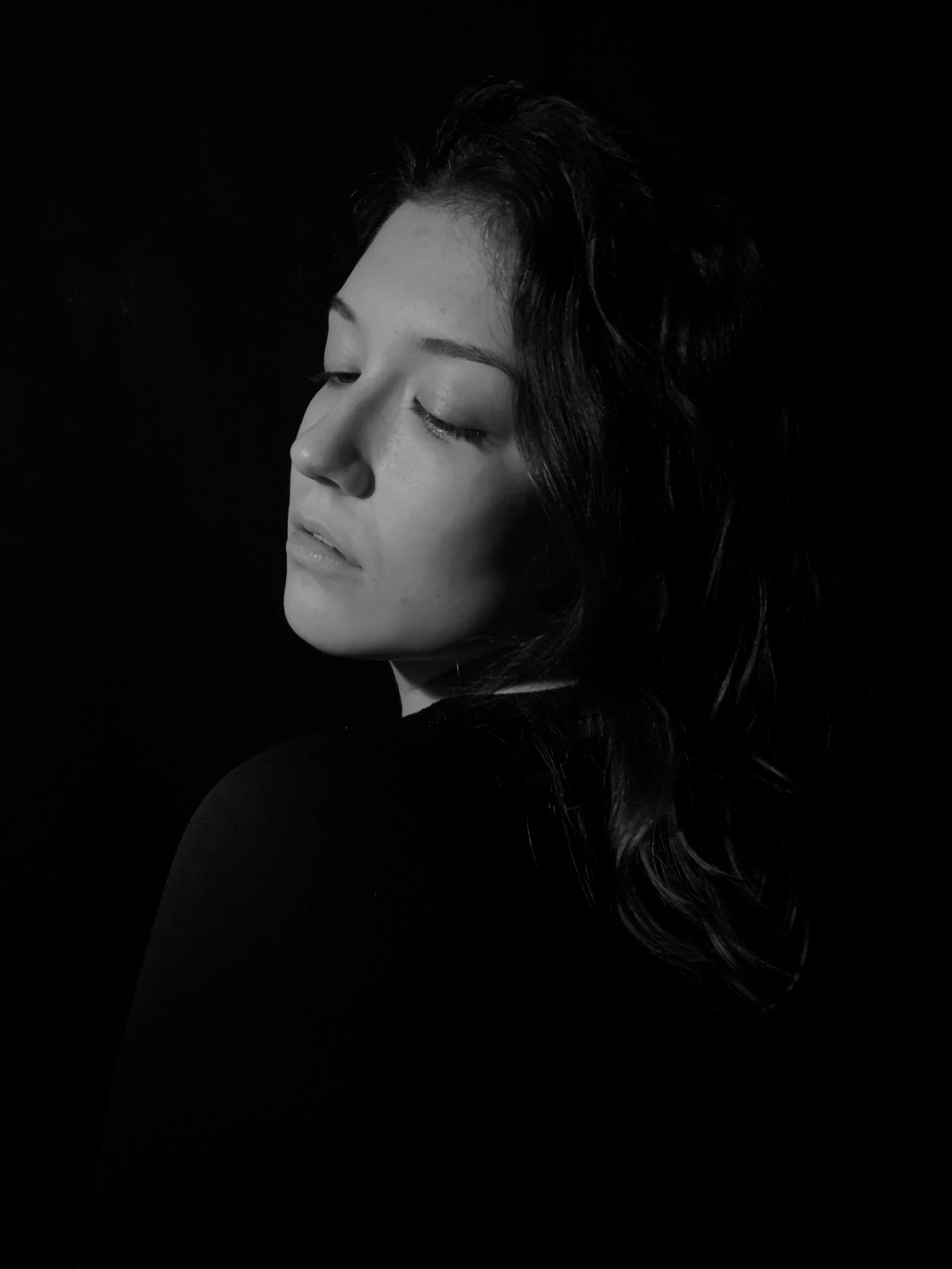 Black and white portrait of woman with closed eyes, looking down, with dark hair against a black background.