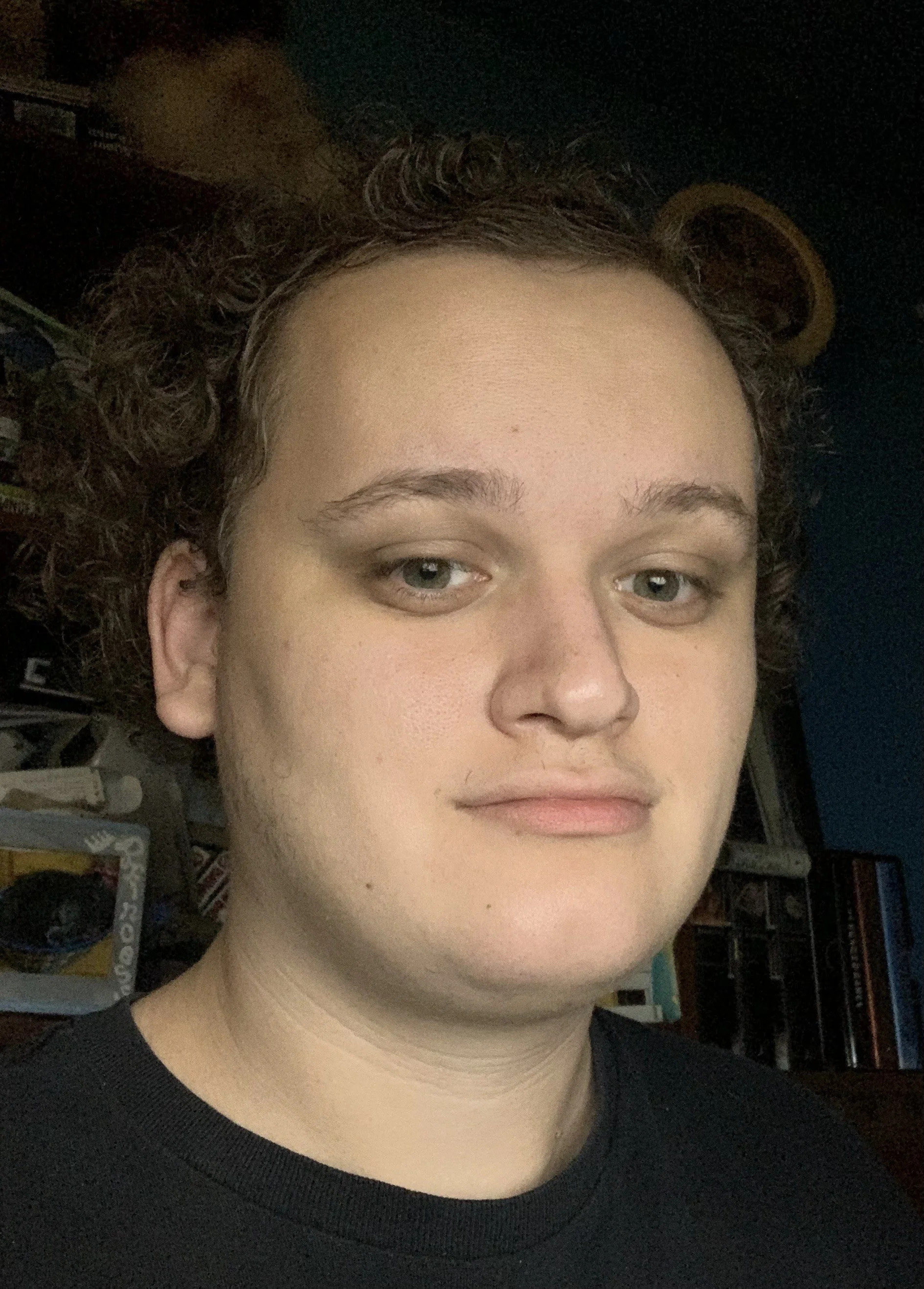 Close-up of a young man with curly brown hair and fair skin, wearing a black shirt, in a dimly-lit room with shelves and books in the background.