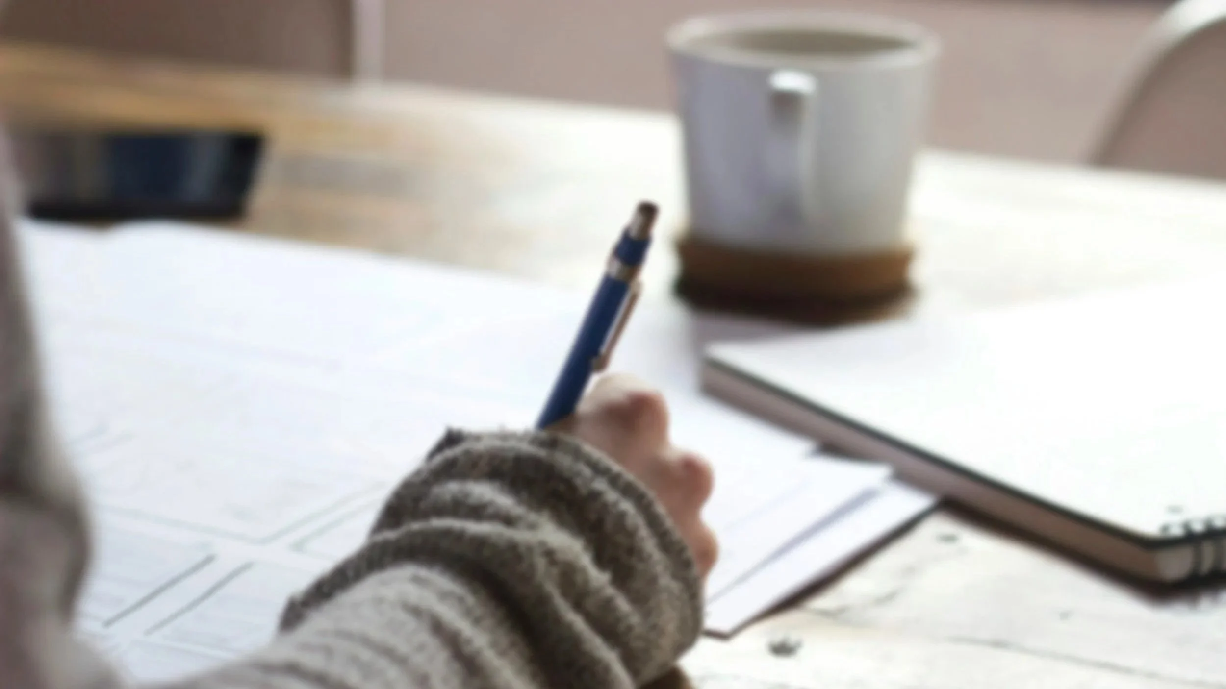 Person writing with a pen on paper, with a laptop, a cup, and a notebook on the table.