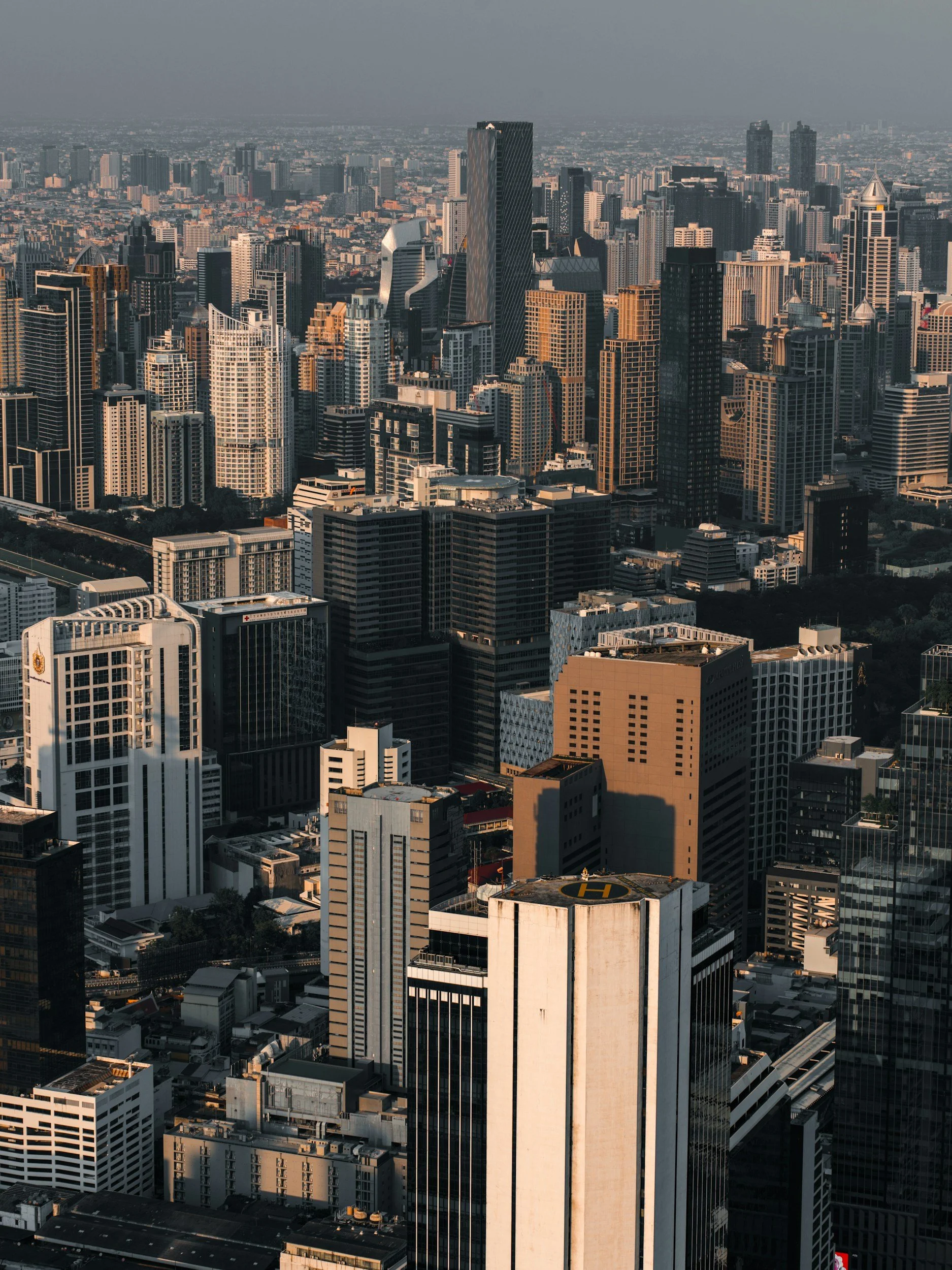 Aerial view of a densely built city with numerous high-rise buildings, skyscrapers, and office towers, some with helipads on the rooftops, under clear daylight.