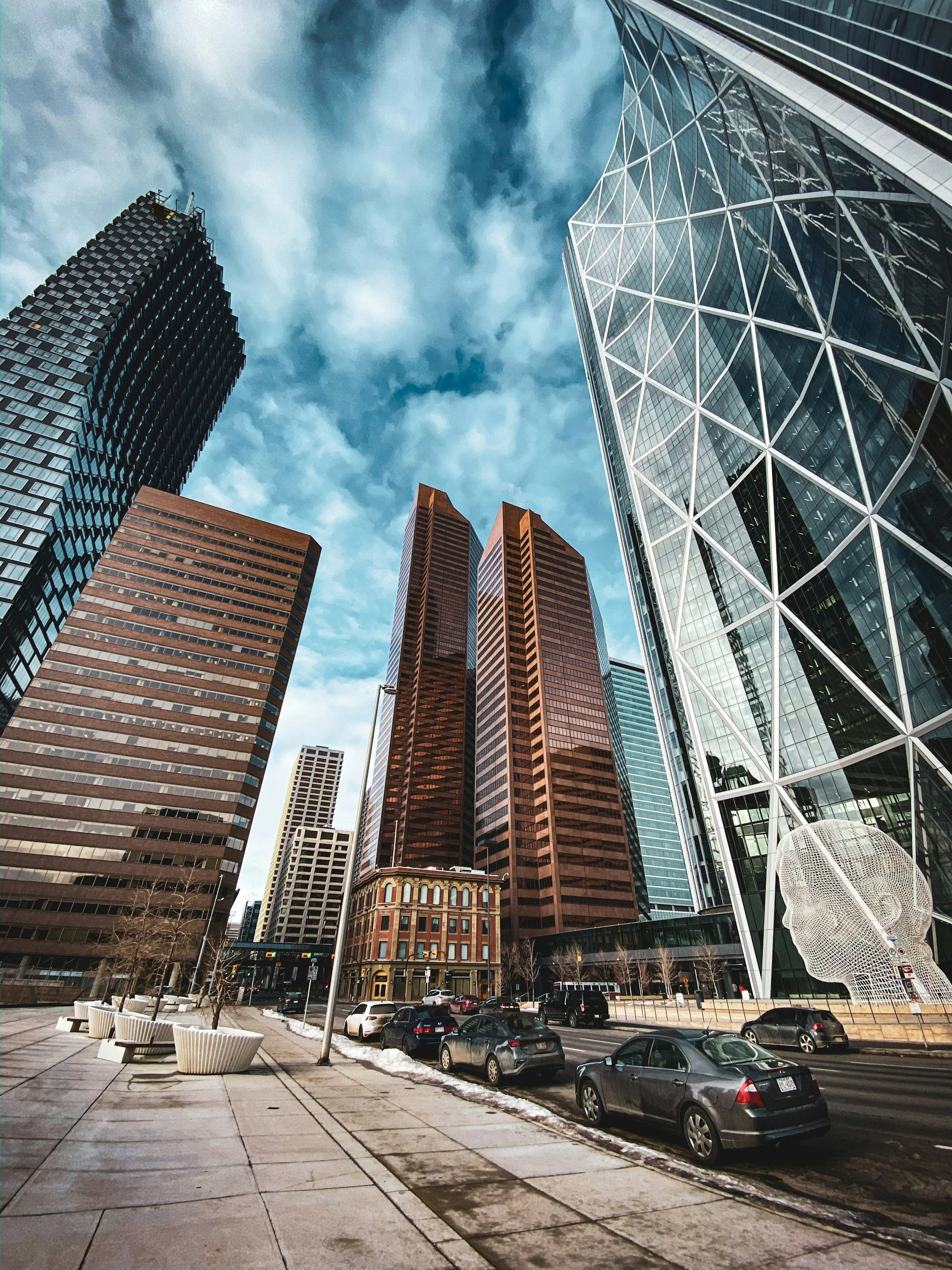 Looking up at tall skyscrapers in a city, with a parking lot and street in the foreground. The buildings have various designs, including glass and brick exteriors, under a partly cloudy sky.