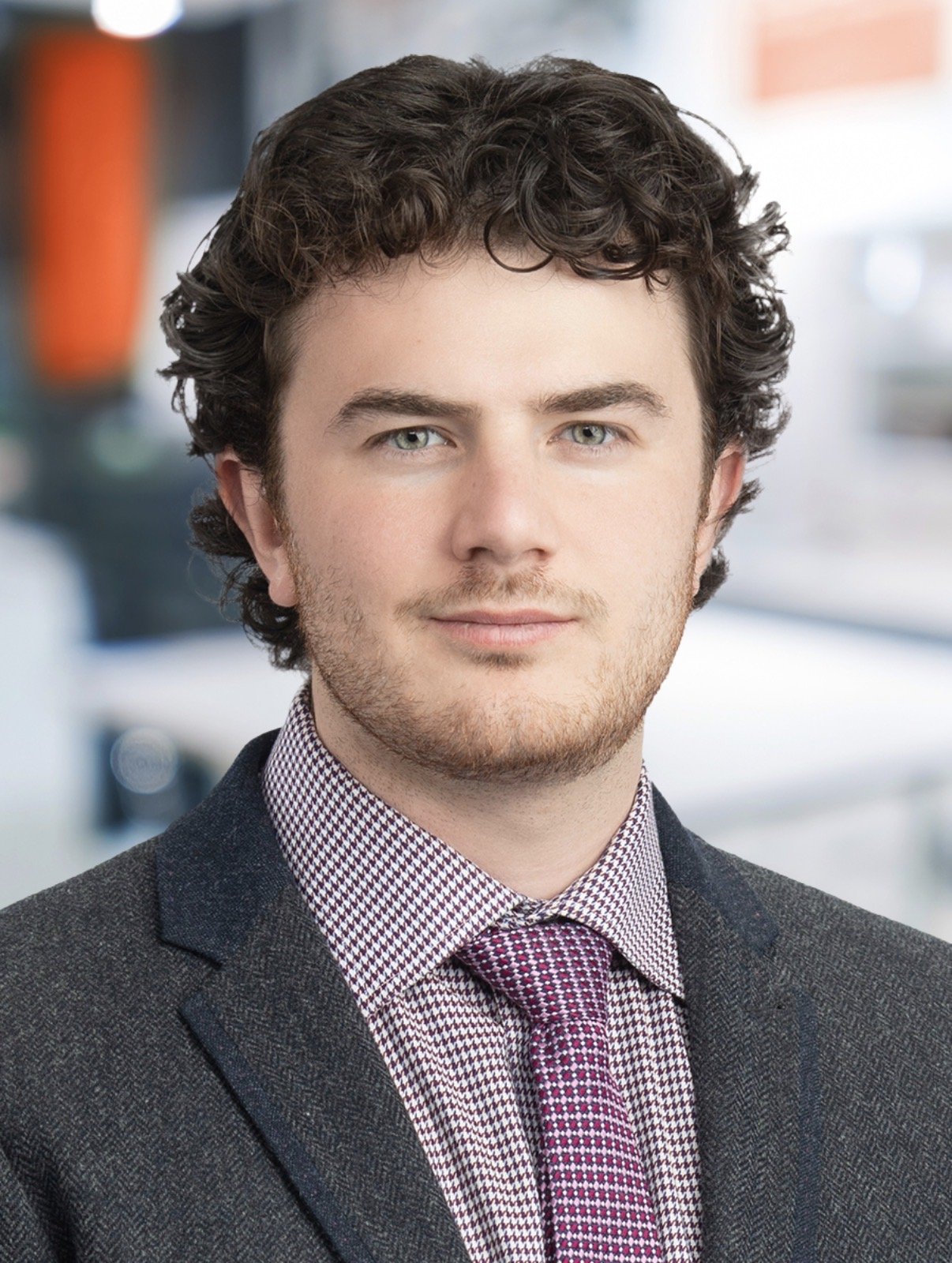 Portrait of a young man with curly dark hair, blue eyes, light skin, wearing a gray suit jacket, checkered shirt, and pink tie in an indoor setting.