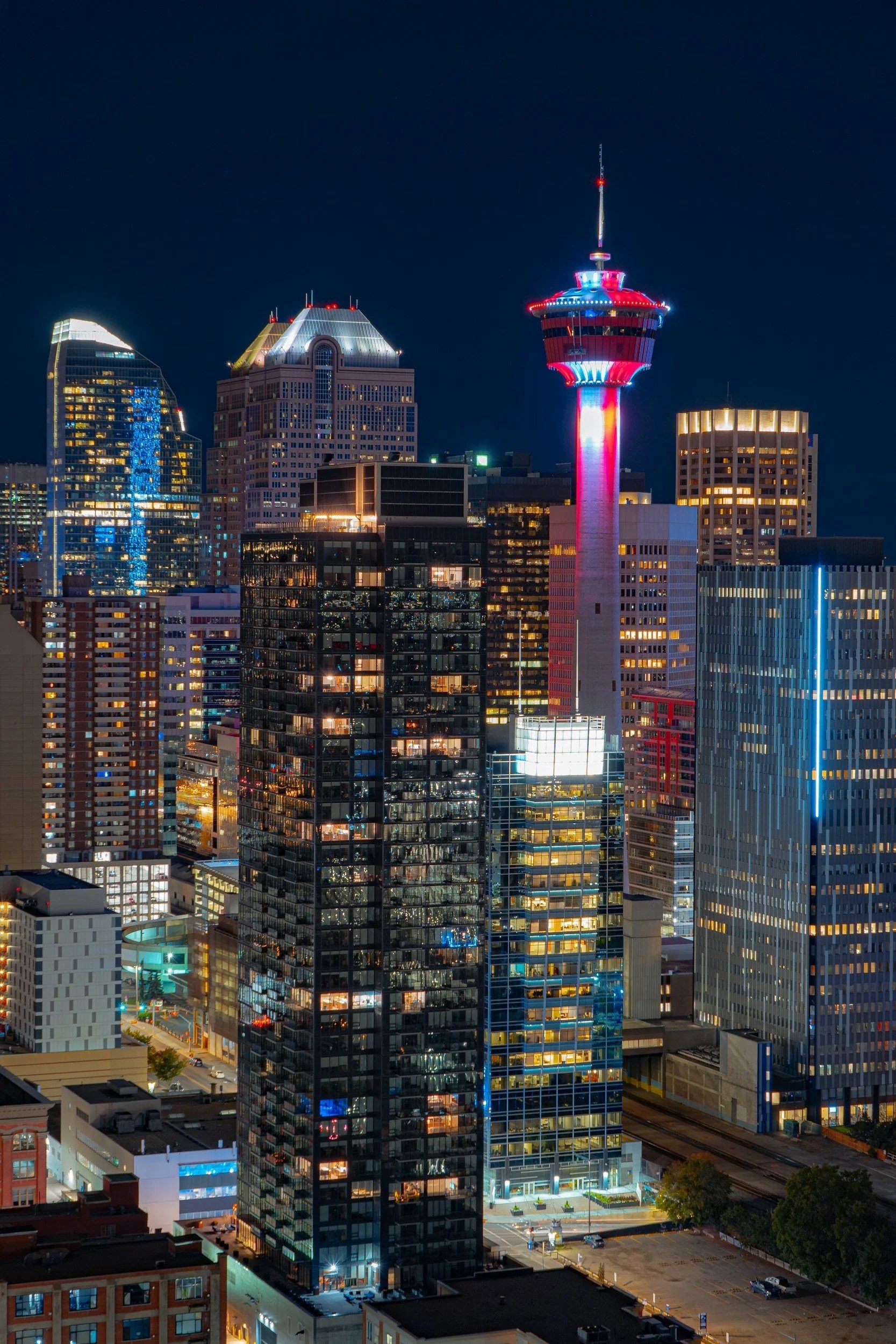 Nighttime cityscape with illuminated skyscrapers, including the Calgary Tower with red, white, and blue lights in downtown Calgary, Alberta.
