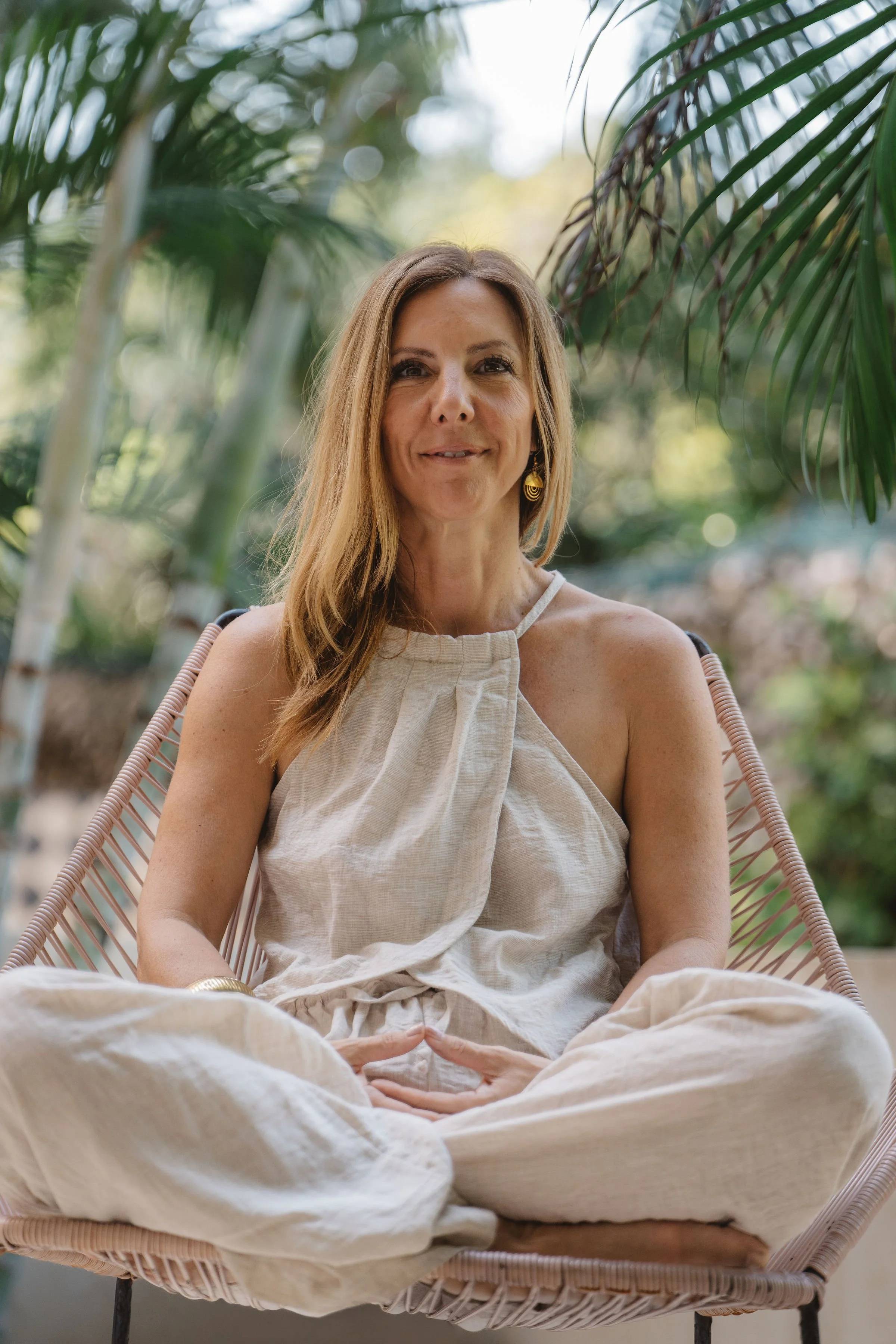 A woman with long, light brown hair sitting cross-legged outdoors in a tan beige dress, framed by green tropical plants.