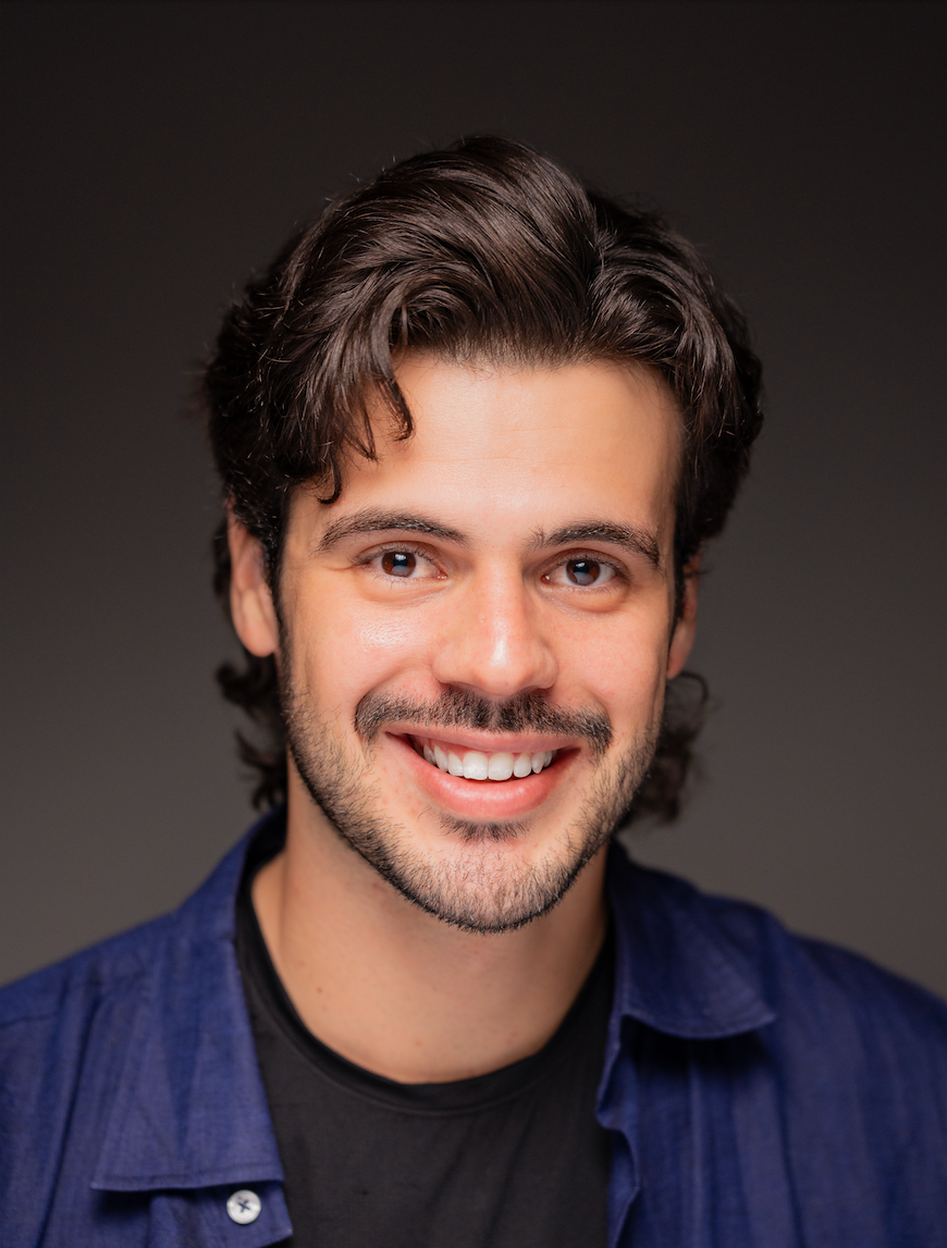 Close-up portrait of a smiling man with dark, curly hair and a beard, wearing a blue shirt and black T-shirt against a dark background.
