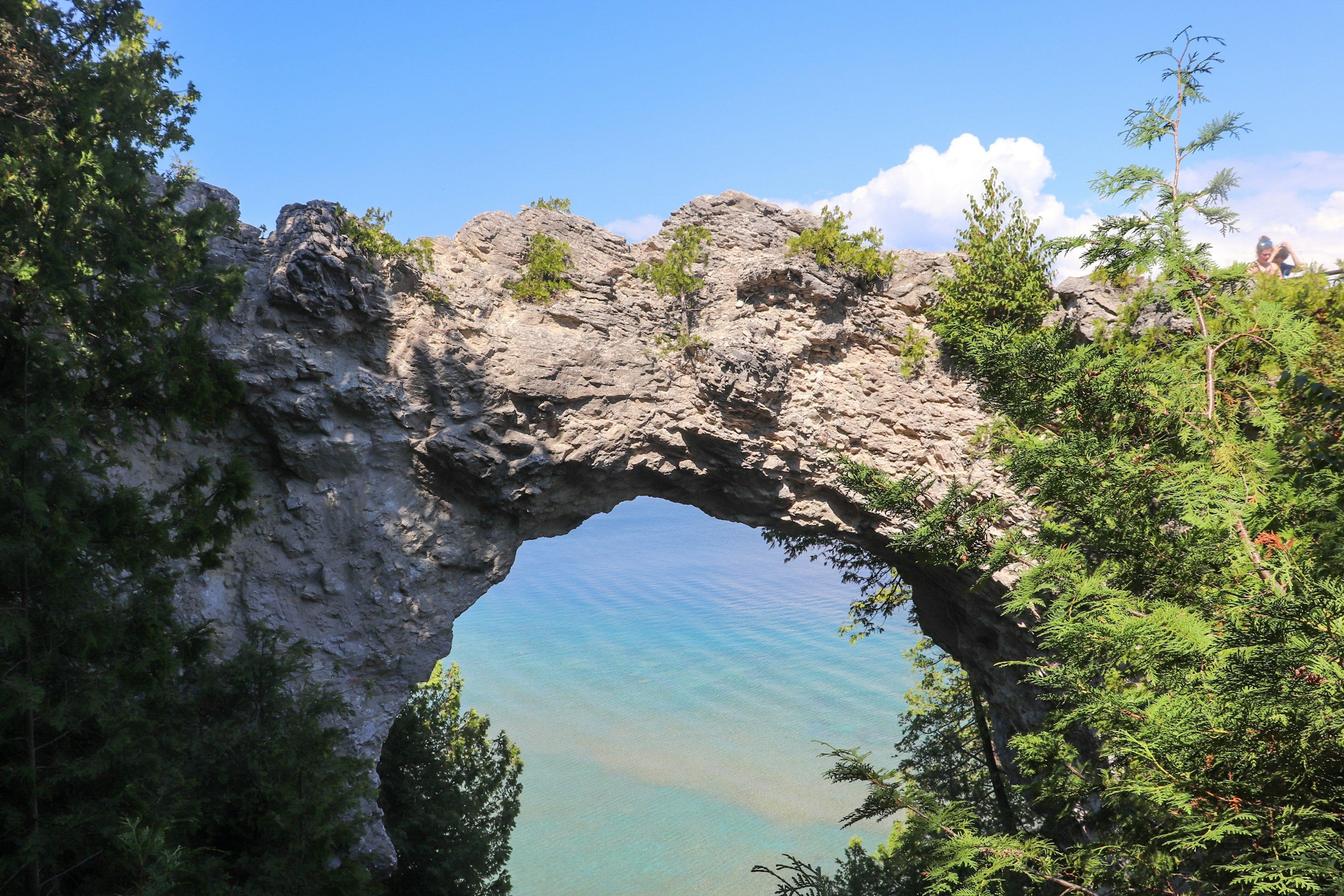 A natural rock arch over a body of water, surrounded by green trees and shrubs, under a blue sky with a few white clouds.