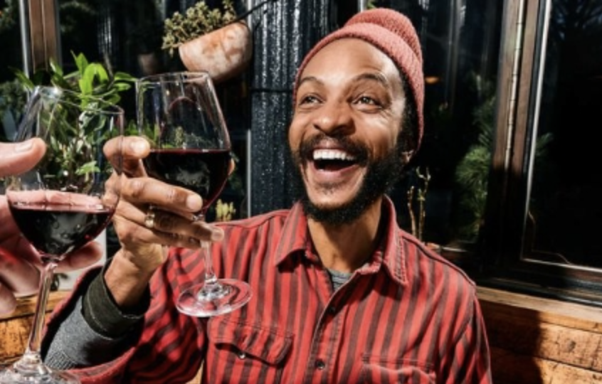 A man with a beard and a pink beanie smiling and clinking glasses of red wine with friends in a cozy, rustic setting.