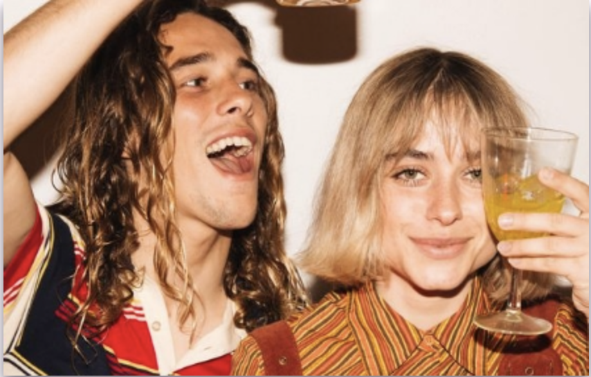Two young adults at a party, one with curly hair and another with straight hair, holding drinks and smiling.