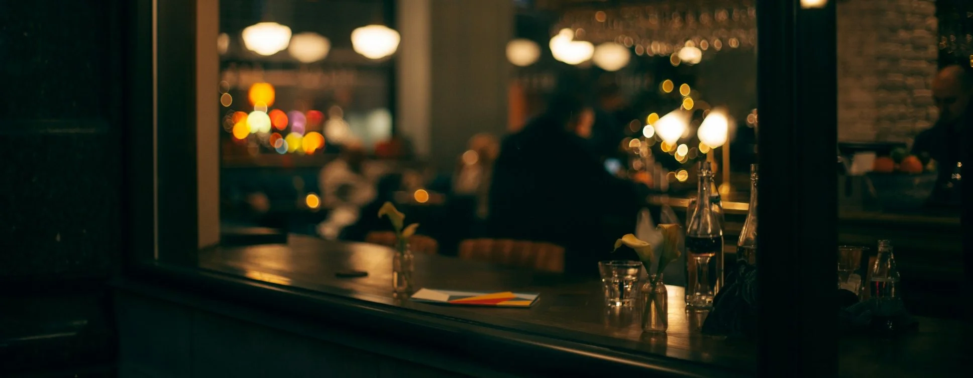 A view through a cafe window showing a dimly lit interior with blurred figures, bottles, and flowers, with city lights in the background.