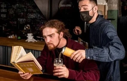 Two men at a bar, one with curly hair and a beard reading a book, the other pouring a drink.