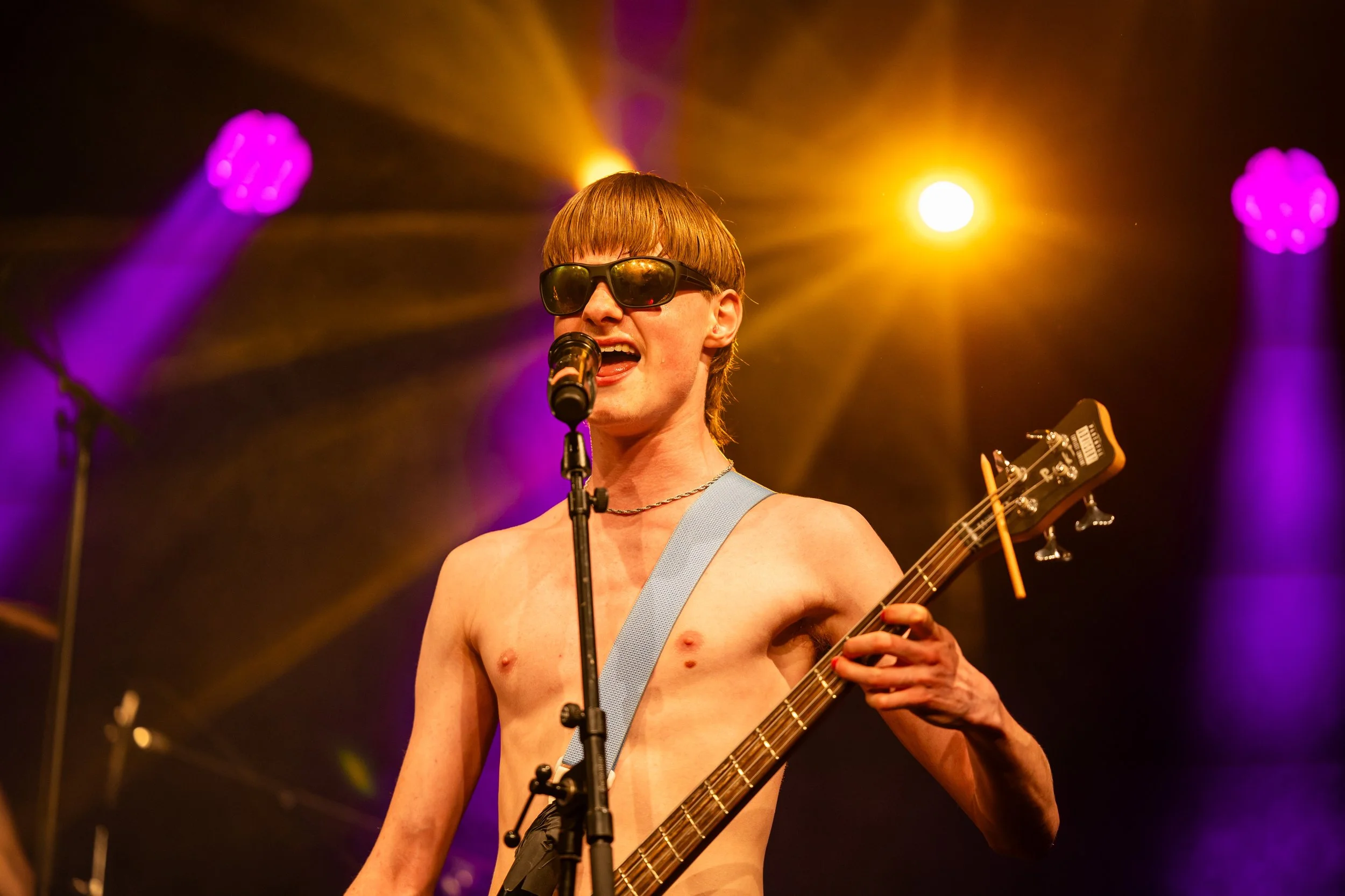 Sæbe from Three Shitting Birds wearing sunglasses singing into a microphone while playing bass on stage with purple and yellow stage lights.