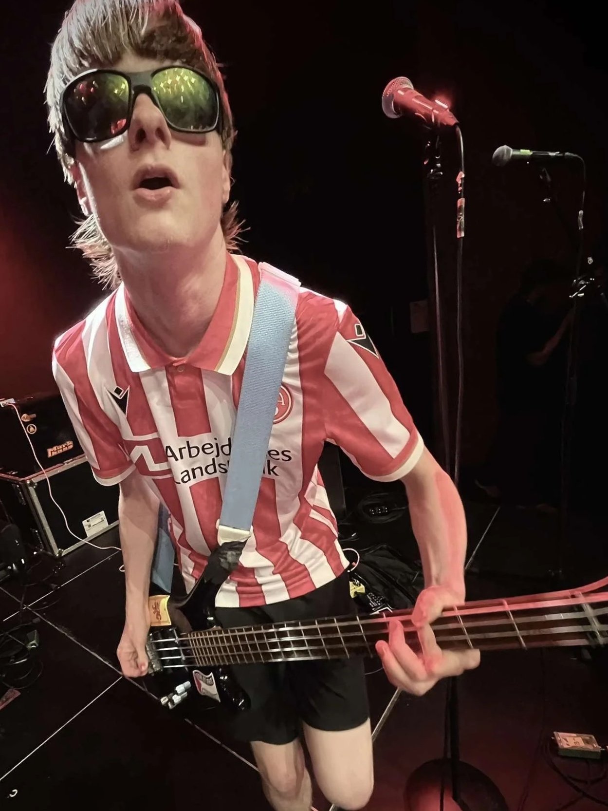 Sæbe form Three Shitting Birds is wearing sunglasses, a red and white striped soccer jersey, and black shorts playing an electric bass guitar on stage with microphones set up nearby.