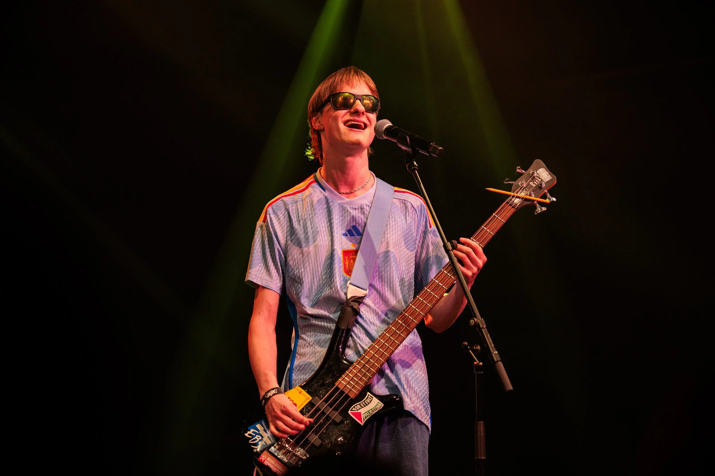 Sæbe from Three Shitting Birds wearing sunglasses and a colorful T-shirt is playing an electric bass guitar and singing into a microphone on stage with green spotlights overhead.