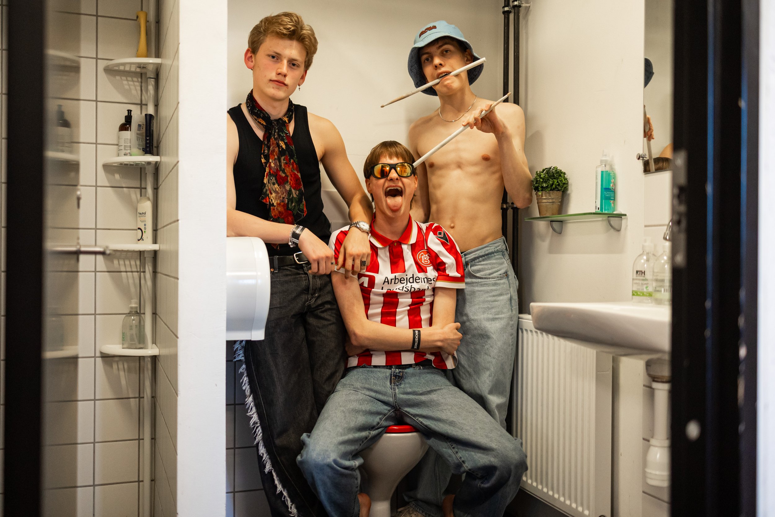Three Shitting Birds in a small bathroom posing playfully for the camera. One is sitting on the toilet with an open-mouthed smile, wearing sunglasses and a red-and-white striped soccer jersey. Two others are shirtless, one wearing a bucket hat and h