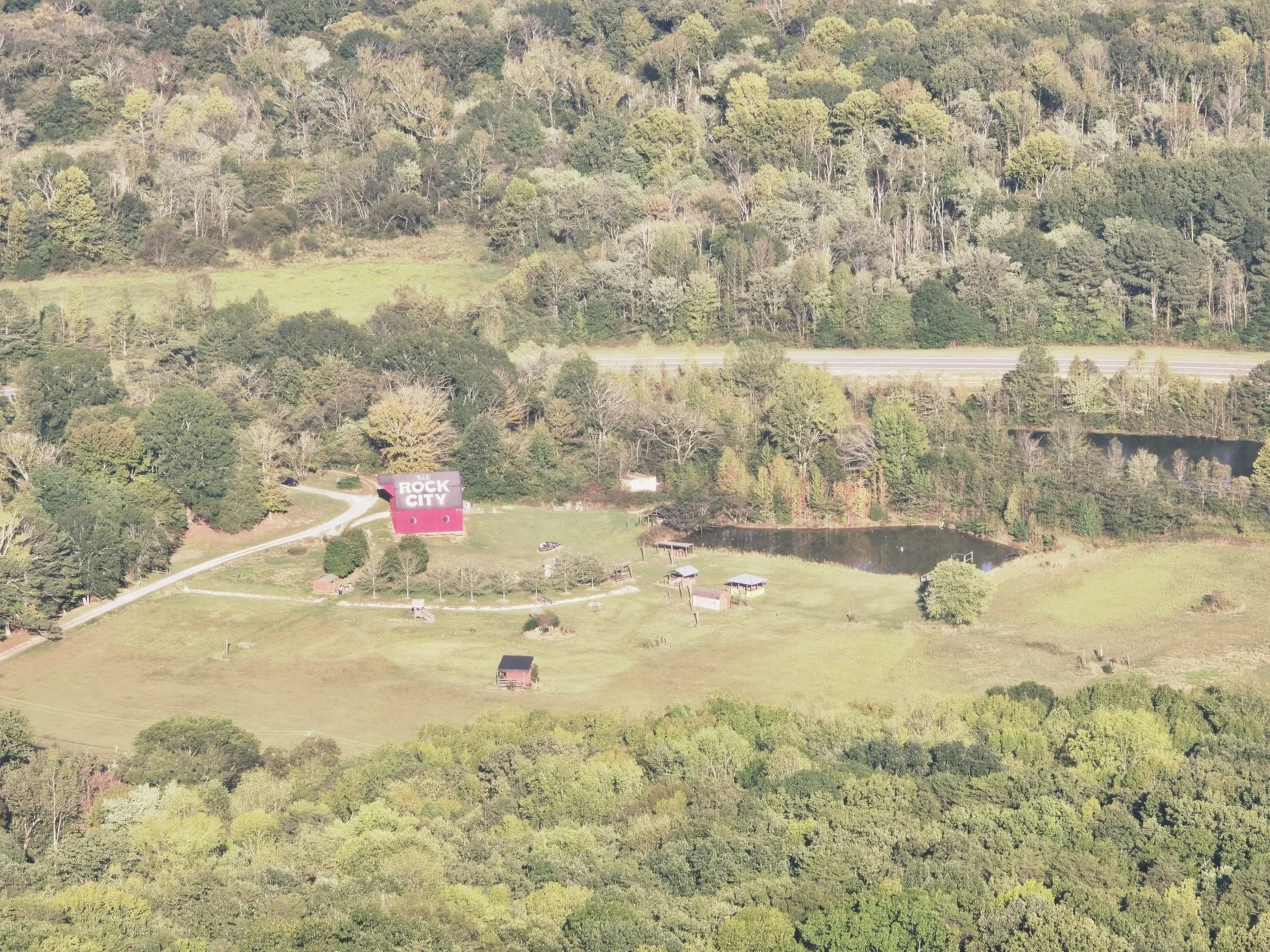 Aerial view of a countryside area with green fields, trees, a small pond, and a building with a pink roof that reads 'Rock City' in white letters.