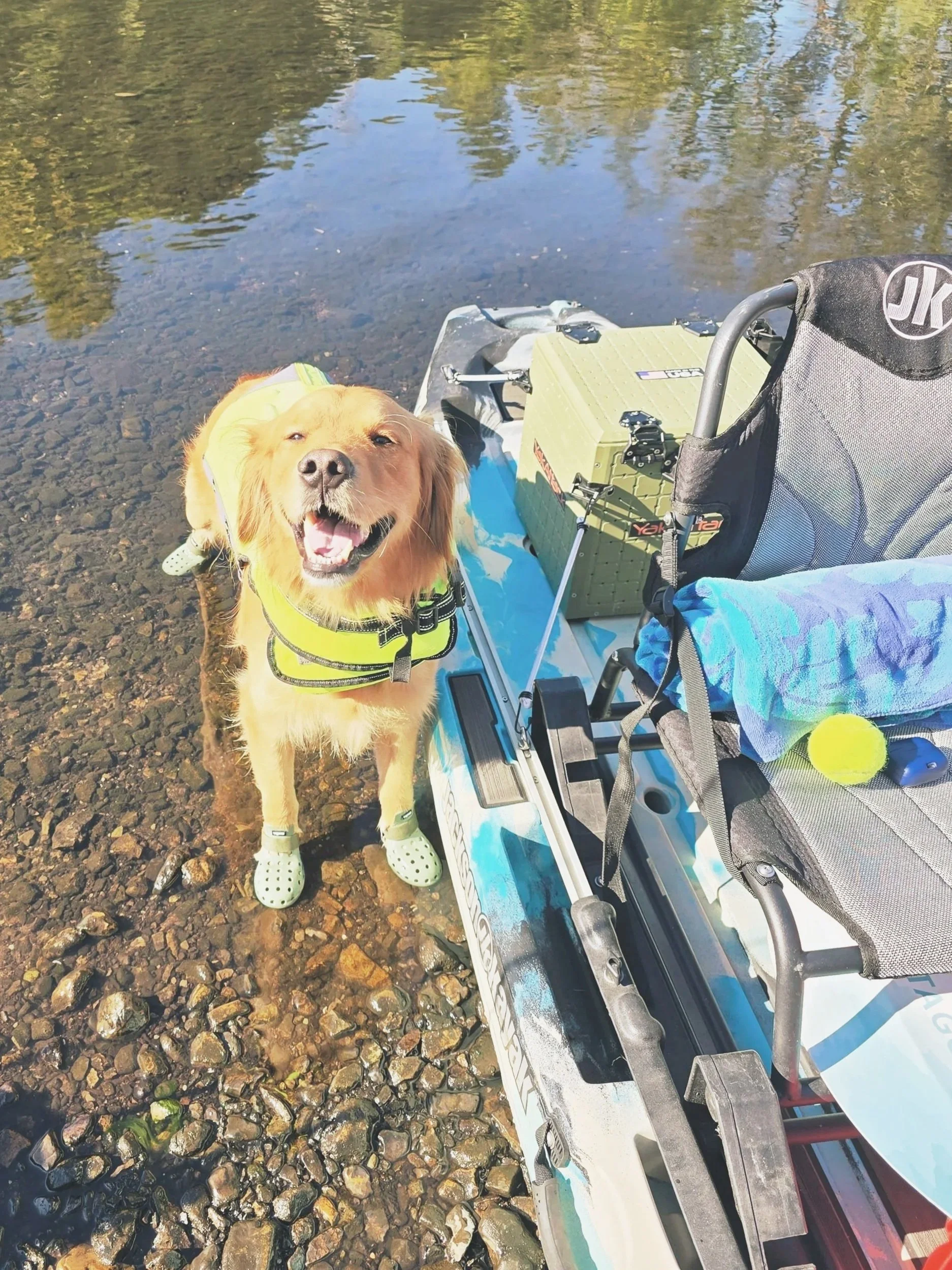 A happy golden retriever wearing a yellow life vest and polka dot water shoes, standing on rocky shore next to a kayak with fishing gear and a tennis ball, near a body of water.