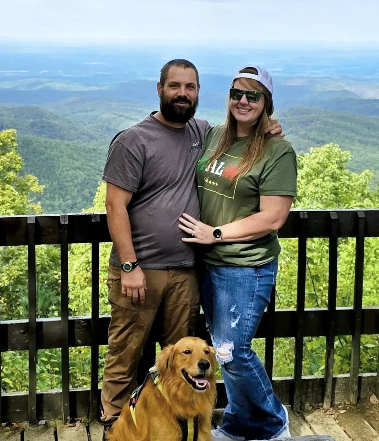 A man and woman standing on a wooden balcony with a scenic mountain view in the background. The man has a beard, is wearing a dark gray t-shirt, brown pants, and a wristwatch. The woman is wearing sunglasses, a baseball cap, a green t-shirt, and ripped jeans. There is a golden retriever dog sitting in front of them, wearing a harness.