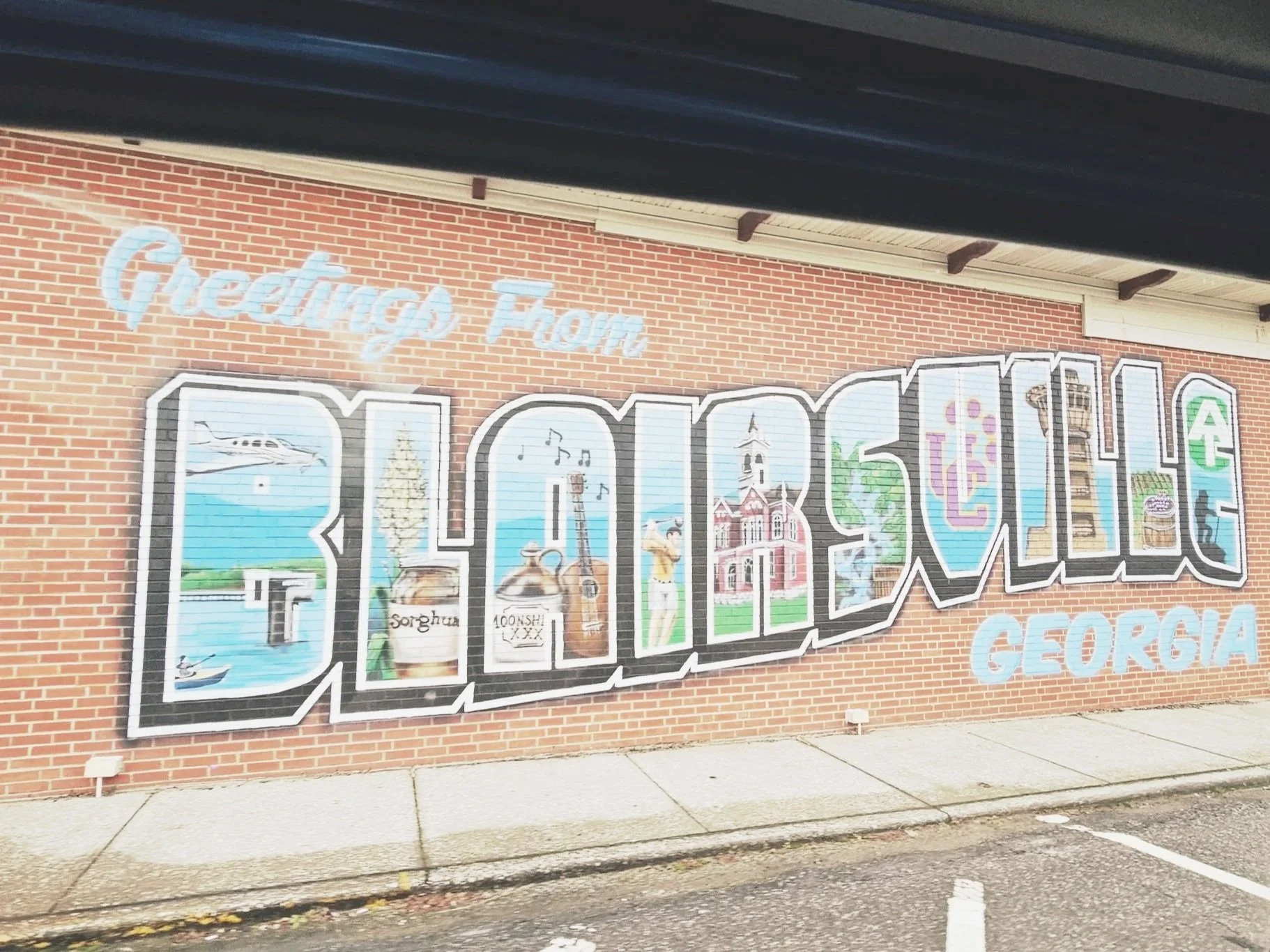 Large colorful mural on a brick wall spelling out 'Brunswick' with images inside the letters, and the words 'Greetings From' above and 'Georgia' below.