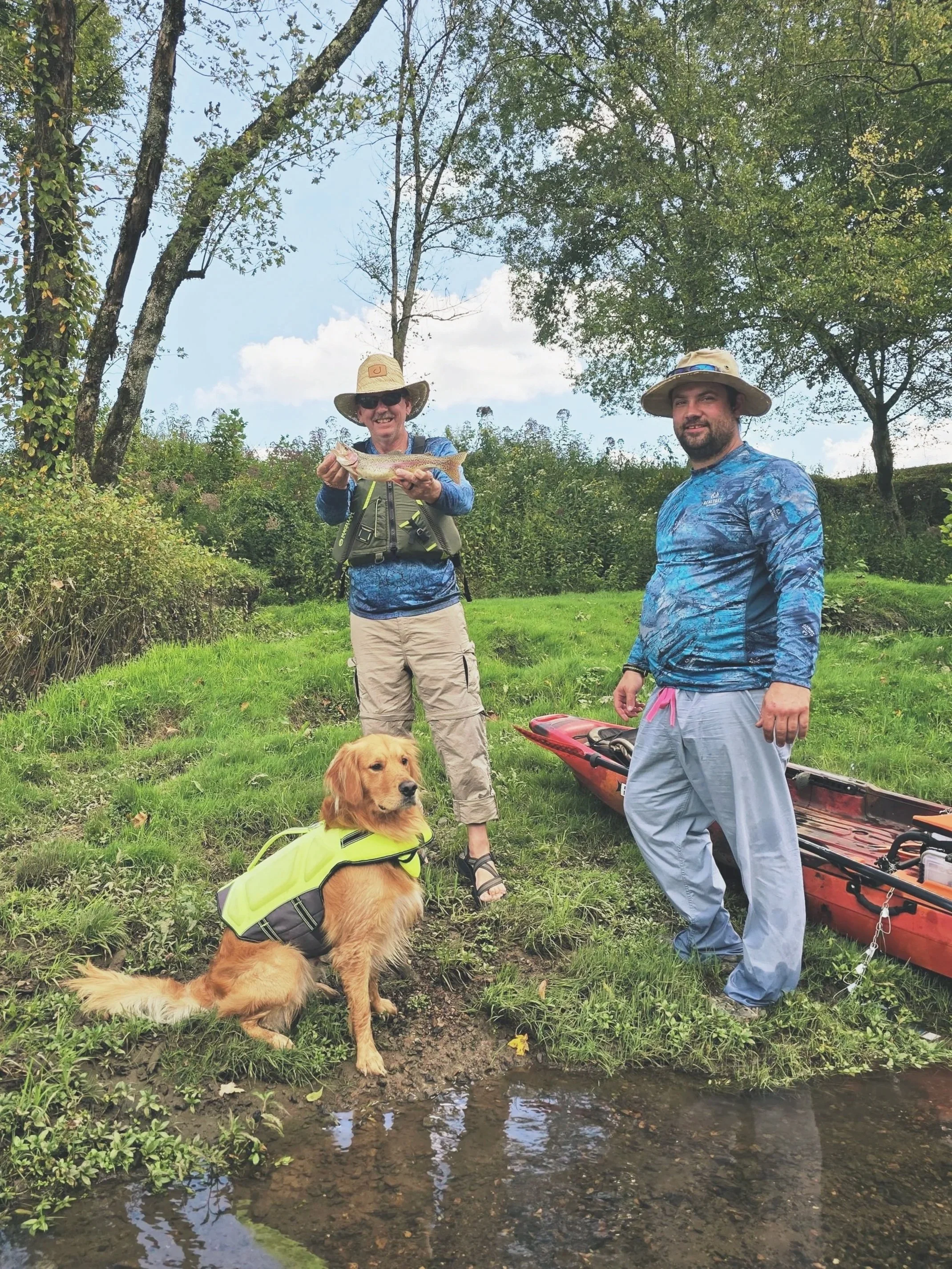 Two men and a dog on a grassy riverbank during a sunny day. One man, holding a fish, is smiling and wearing a straw hat, sunglasses, and outdoor gear. The other man, standing nearby, wears a wide-brimmed hat, a blue athletic shirt, and light-colored pants. A golden retriever wearing a yellow life vest sits in front of them, near the water. A kayak is on the ground next to the second man.