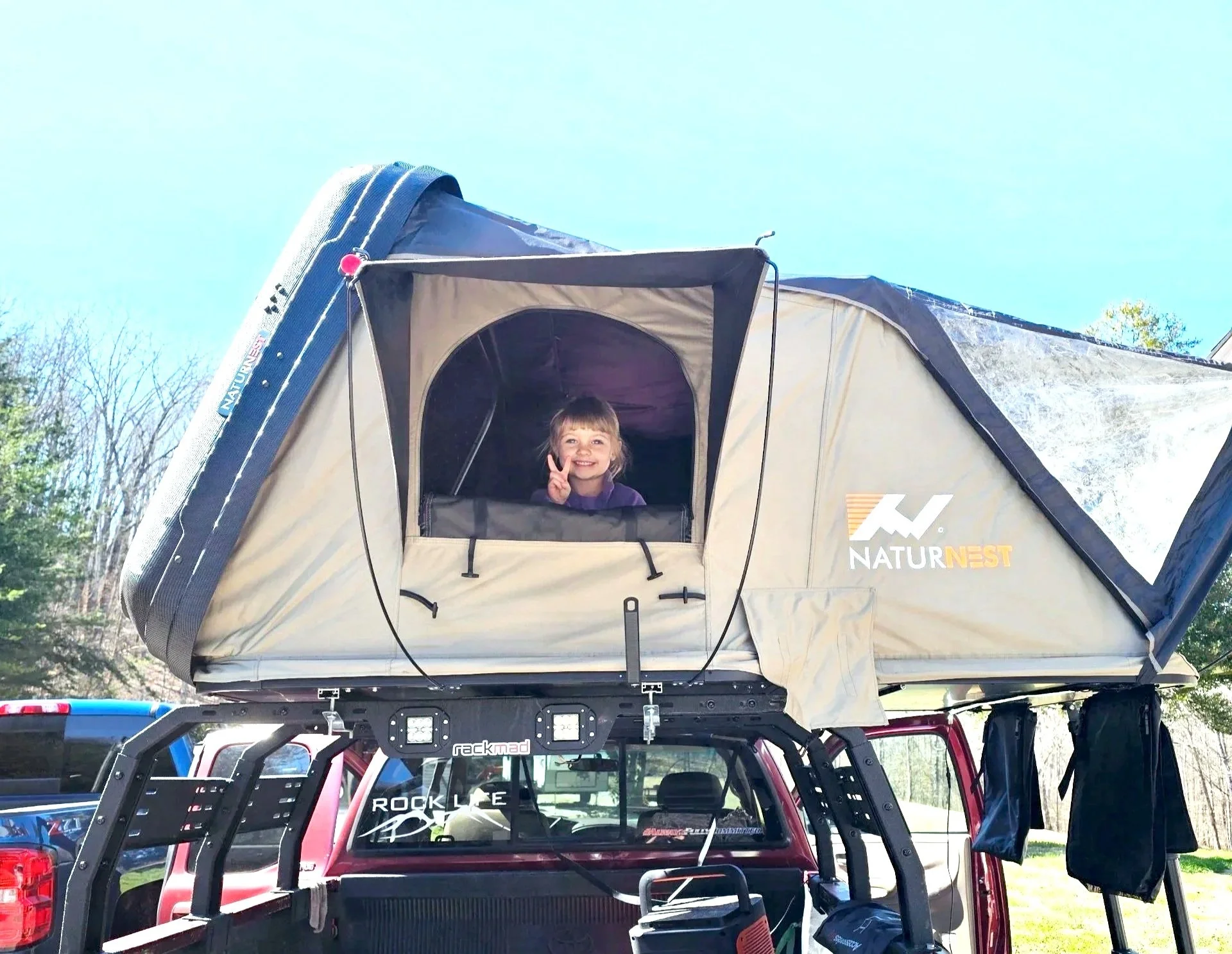 A young girl smiling and making a peace sign from a rooftop tent mounted on a red truck, with trees and a clear blue sky in the background.
