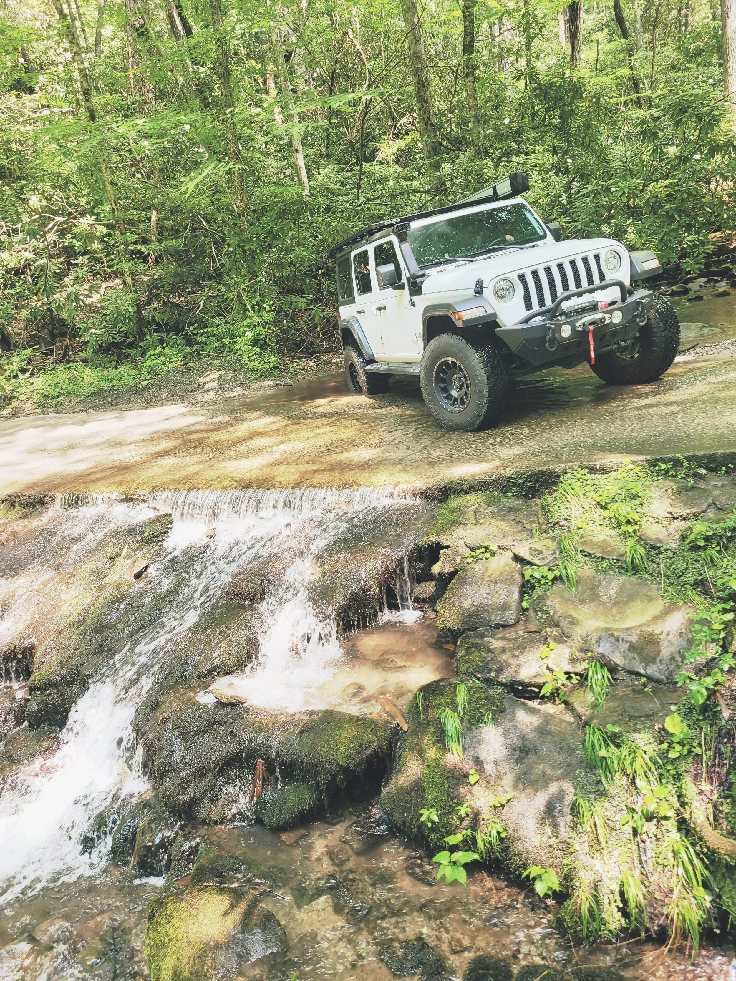 A white Jeep Wrangler off-road vehicle parked on a shallow rocky stream with a forest background.