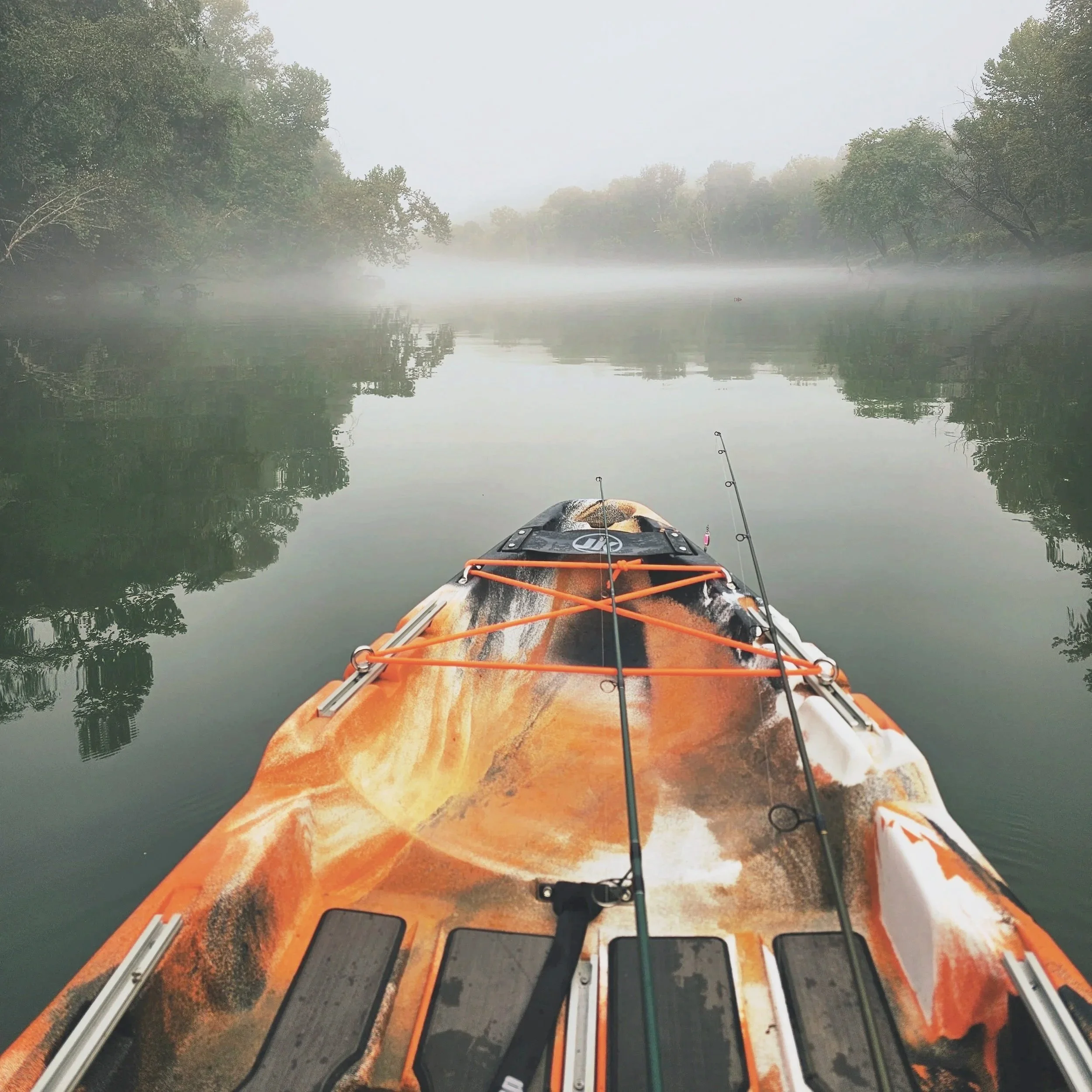 View from a kayak on a calm river with foggy, tree-lined banks, and fishing rods on the kayak