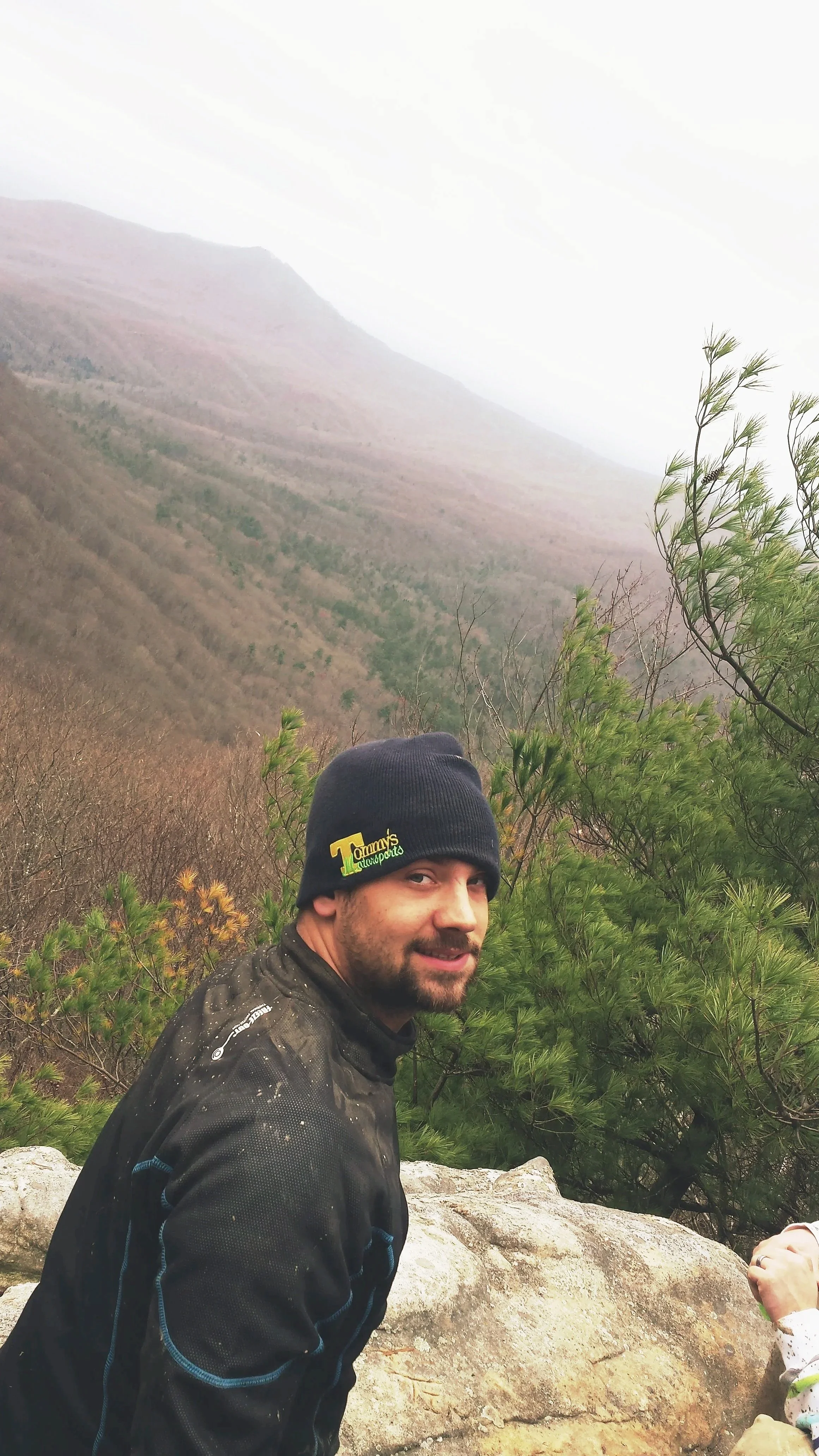 A man with a beard wearing a black beanie and black jacket, smiling at the camera, with a scenic mountainous background and green foliage.