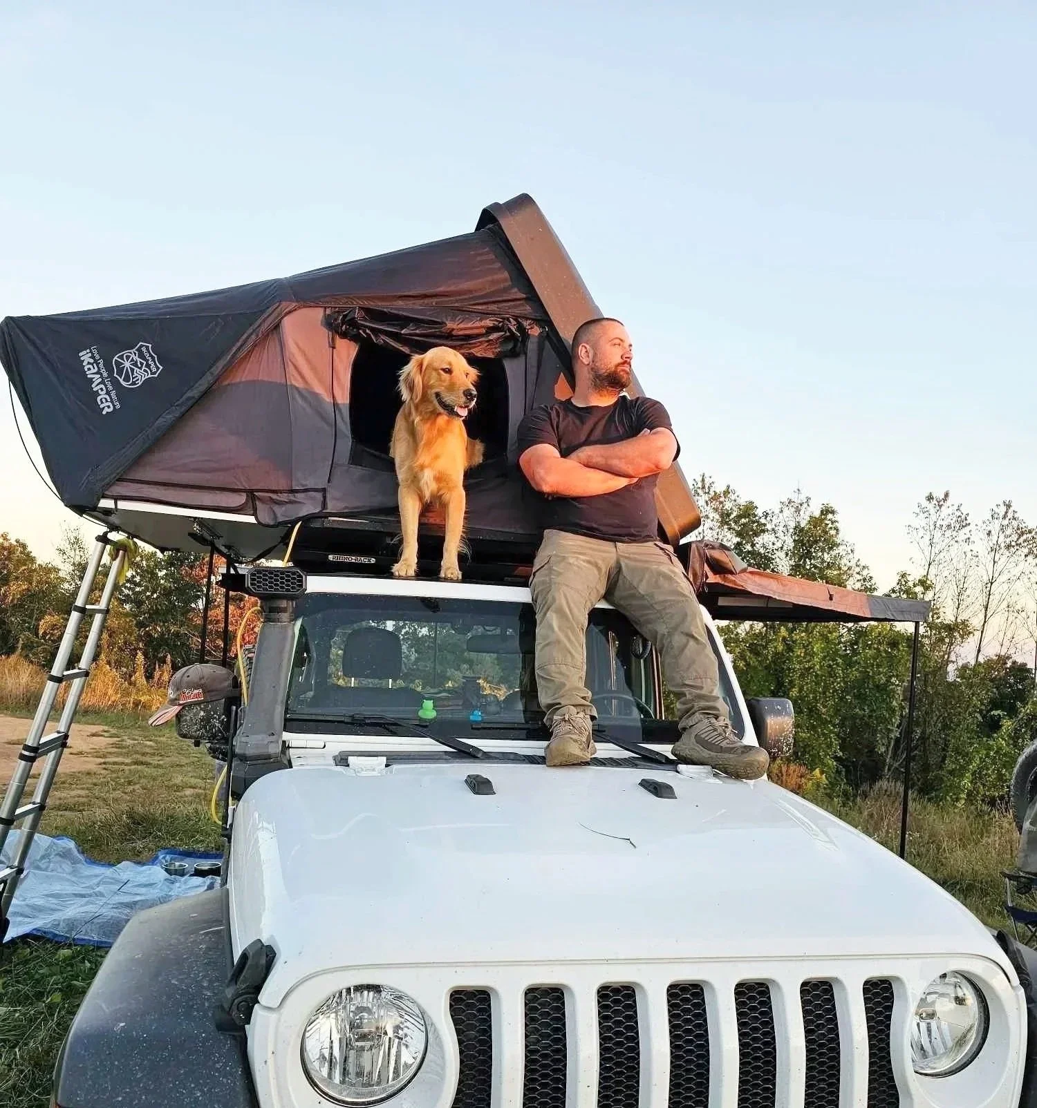 A man and a dog sitting on the roof of a white vehicle with a rooftop tent, in a rural outdoor setting.